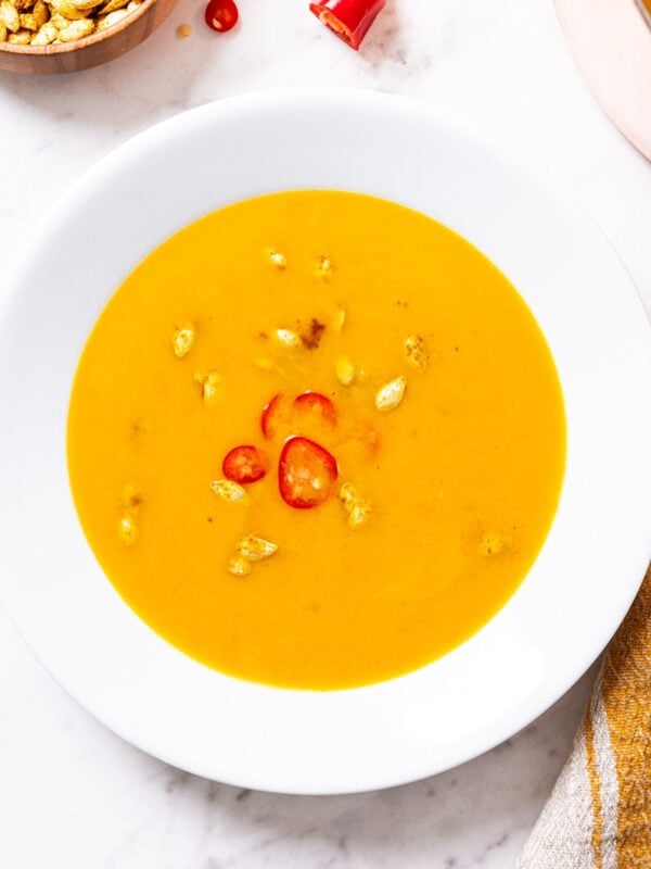 White bowl filled with serving of butternut squash soup with squash seeds and slices of red pepper floating on surface of soup.