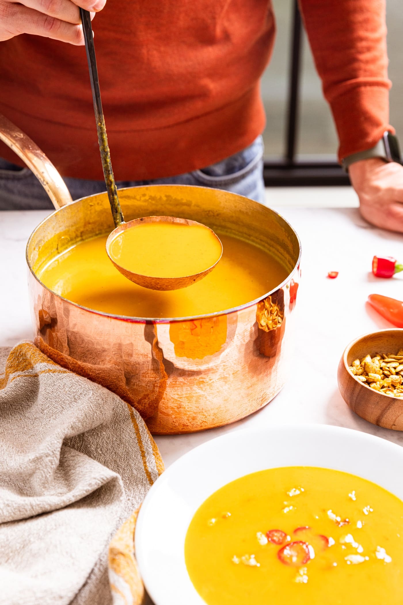 Hand holding copper colored ladle scooping out serving from copper stockpot sitting on marble surface with serving of soup already in white bowl.