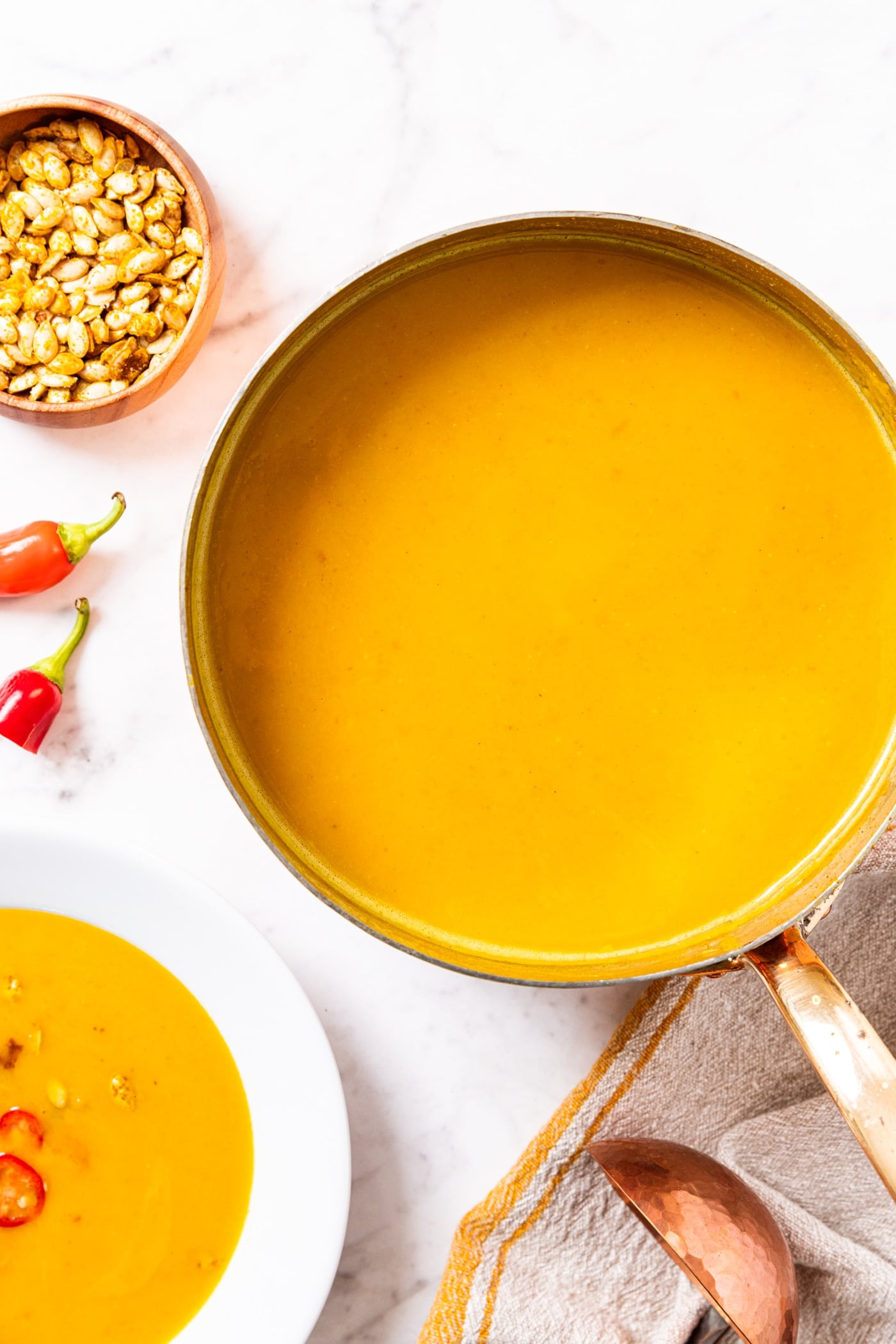 Copper pot filled with yellow colored squash soup sitting on marble surface with white bowl filled with soup and squash seeds in wooden bowl.