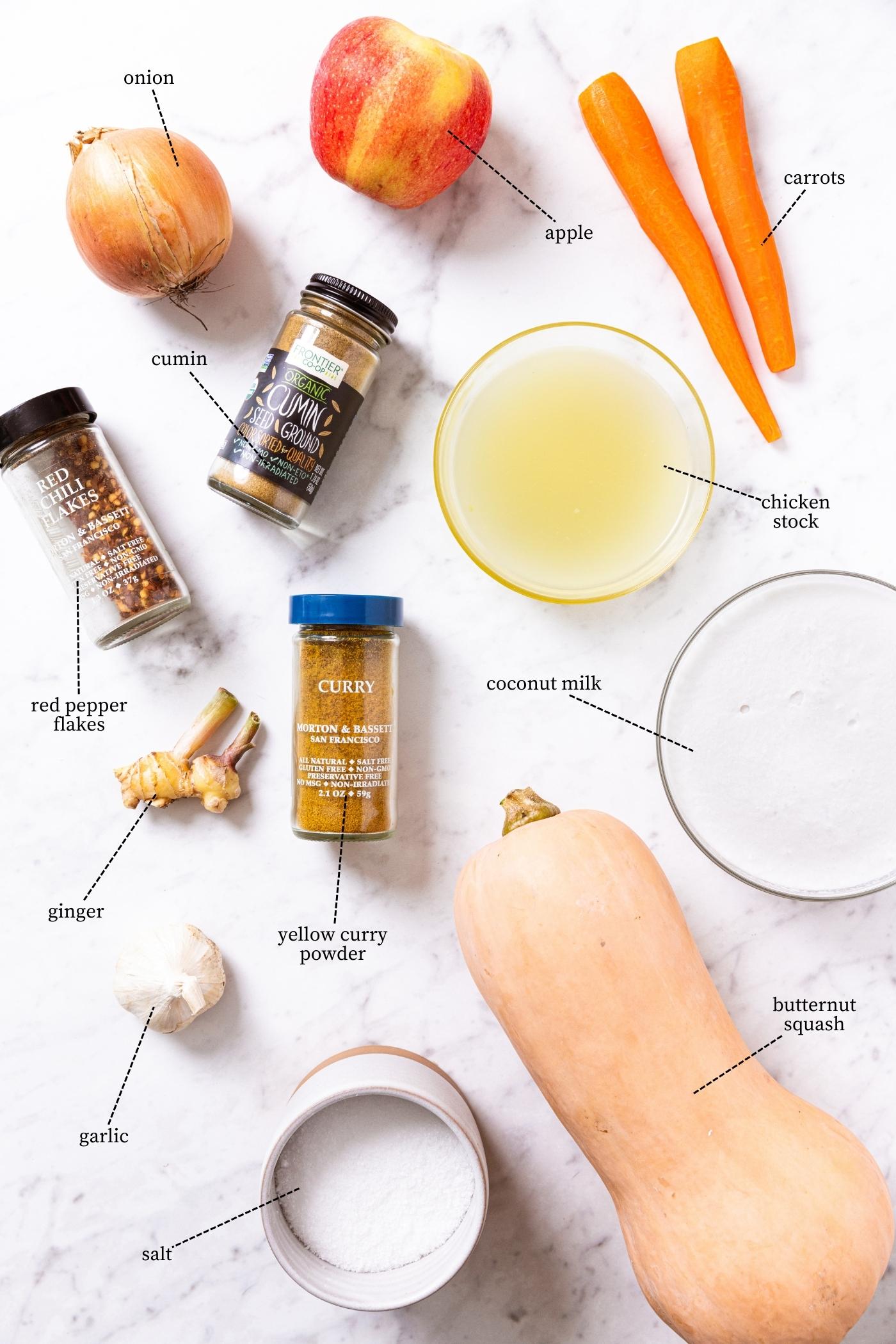 Marble surface filled with all the ingredients needed to prepare butternut squash soup including squash, carrots, onion, apple, spices, salt, and more.