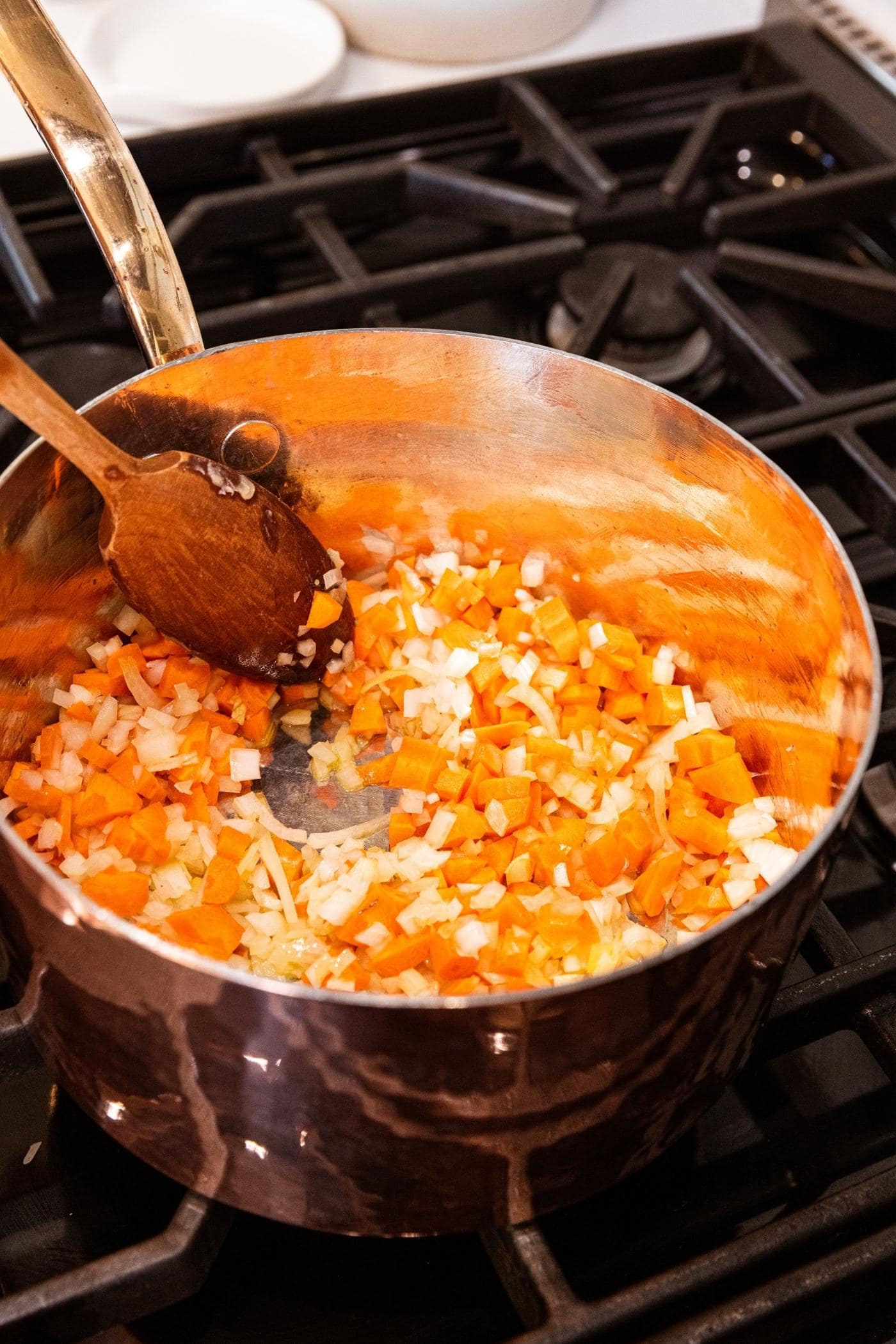 Wooden spatula mixing together onion and carrot mixture on the stovetop in copper pot.