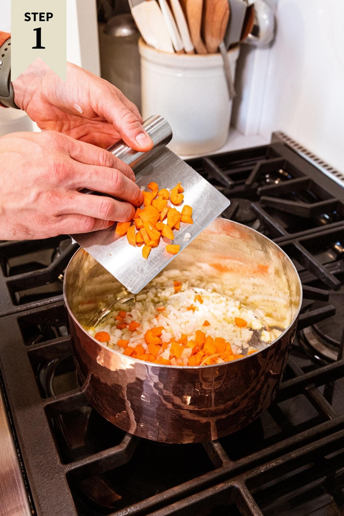 Hand holding metal bench scraper putting diced carrot pieces into copper pot on stove with more carrots and onion already heating.