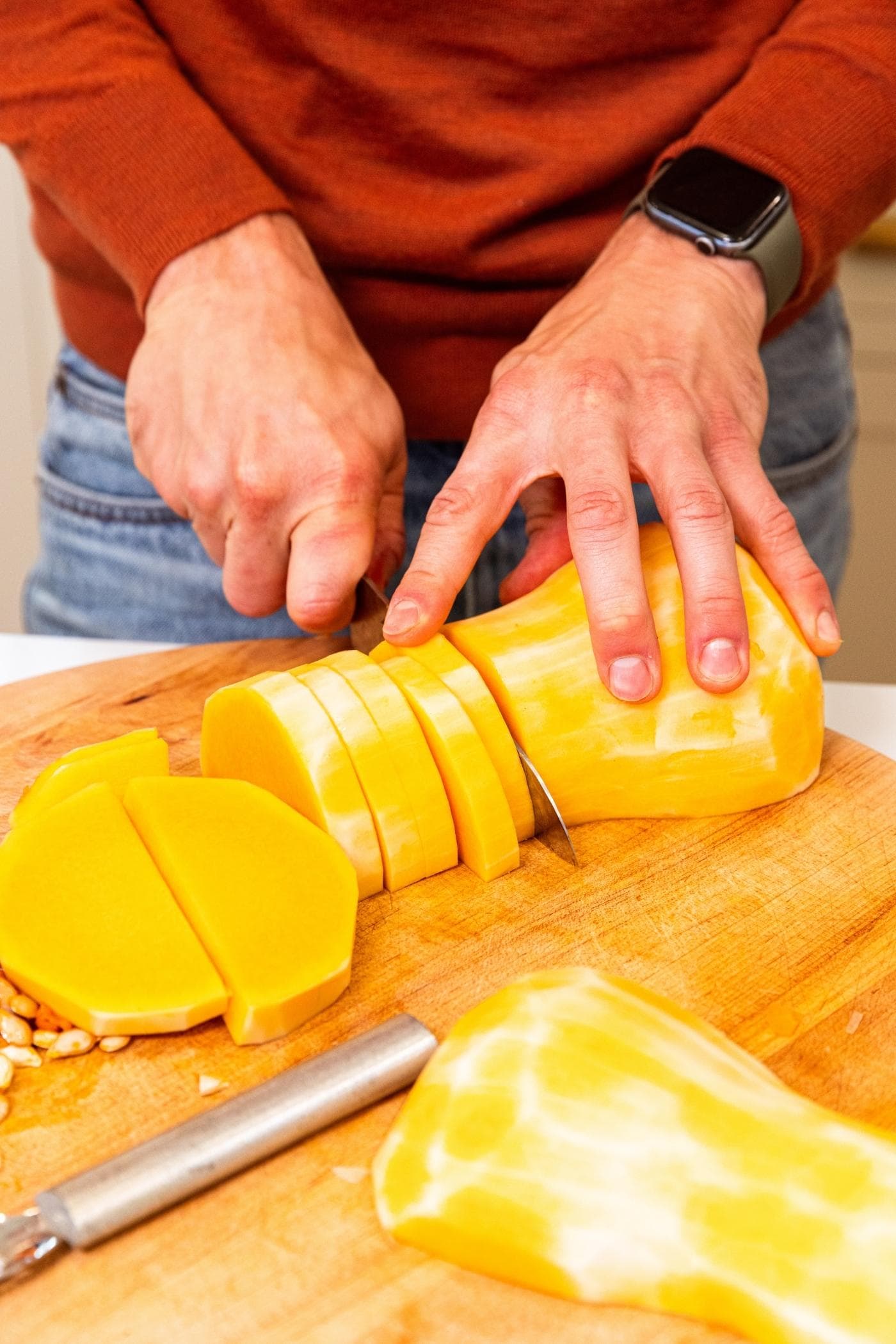 Hand slicing butternut squash into slices before dicing on wood cutting board.