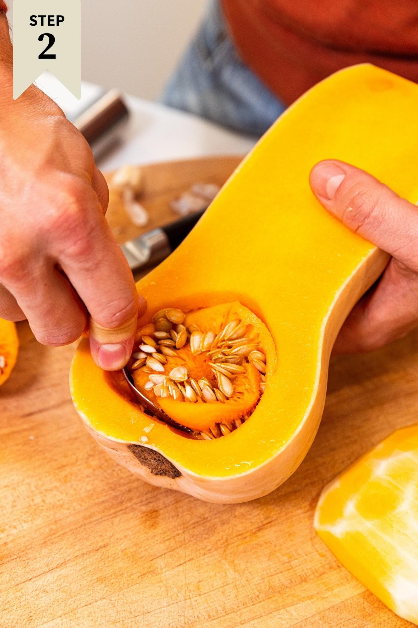 Hand using spoon to scoop out seeds from a butternut squash on a wood cutting board.