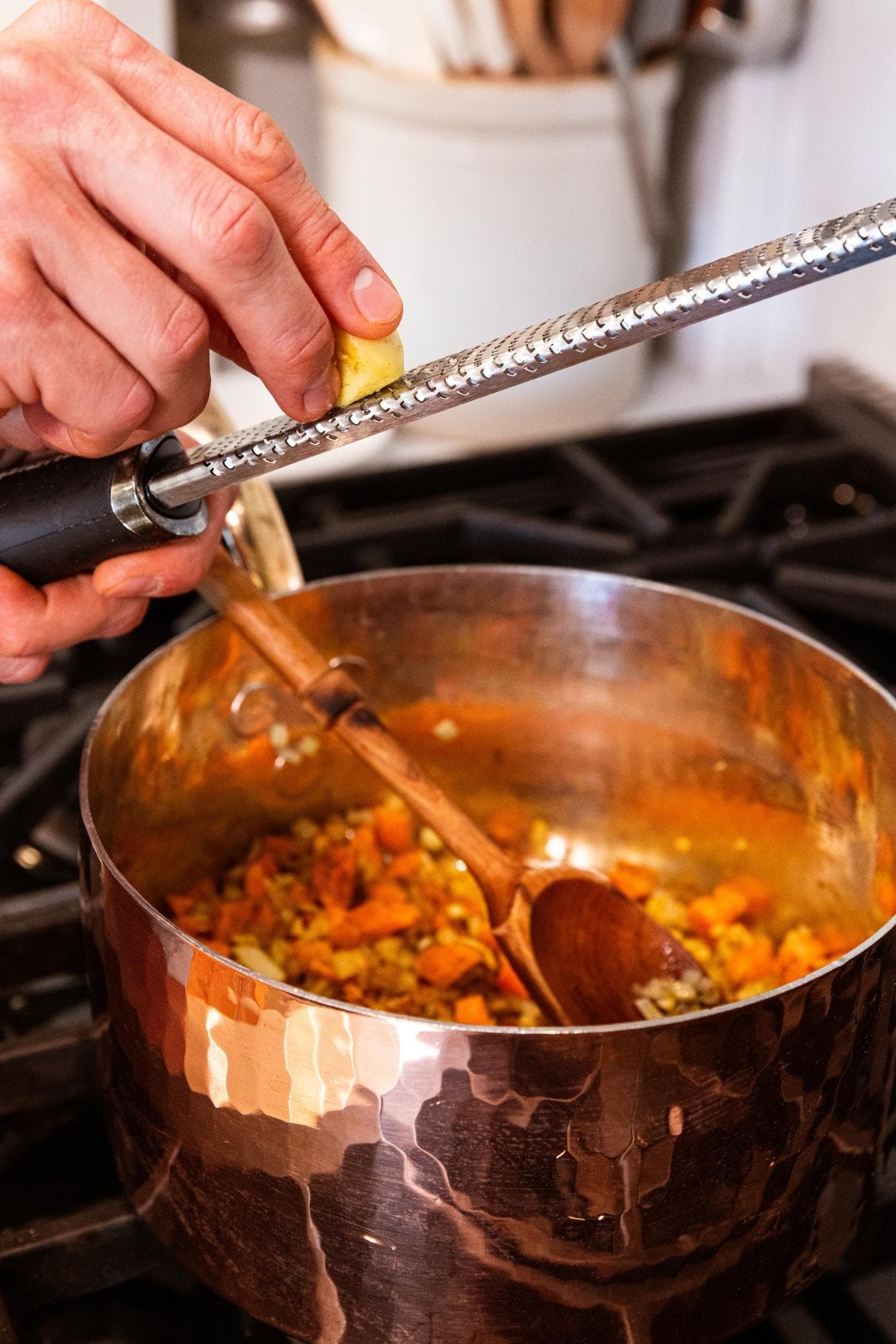 Hand using grater to grate fresh ginger into copper pot sitting on stove with spices and carrots and onion inside.