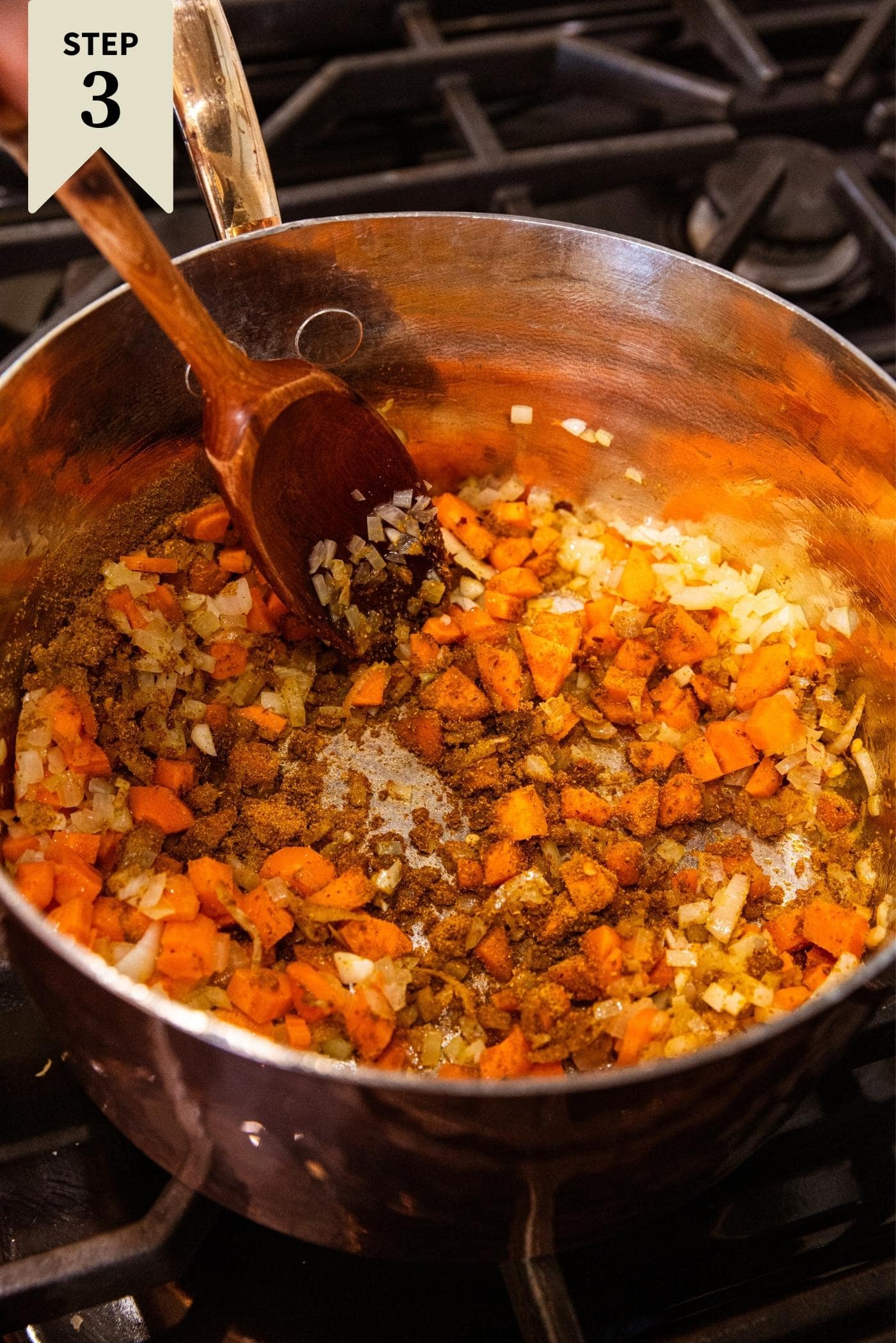 Wooden spatula in copper pot mixing in spices with carrots and onions.