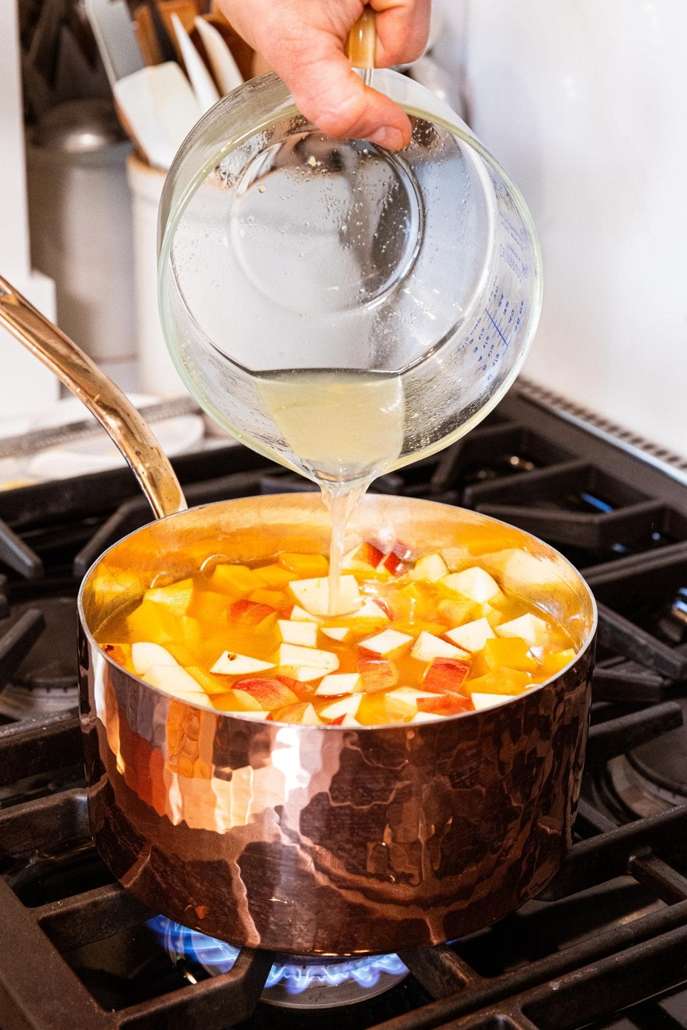 Hand pouring chicken stock from glass measuring cup into copper pot filled with diced butternut squash and apple.