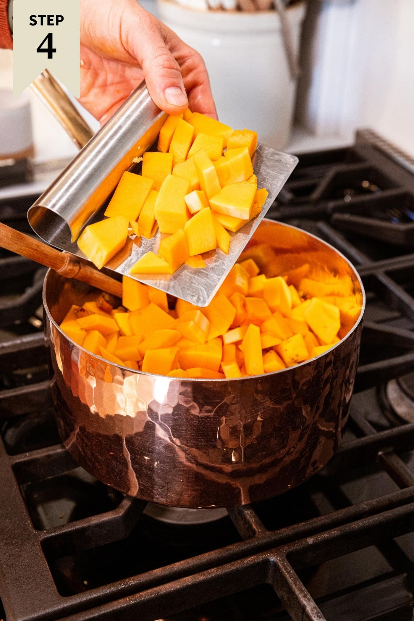 Hand using metal bench scraper to put chunks of chopped butternut squash into copper pot on stovetop already filled with carrots and onions.