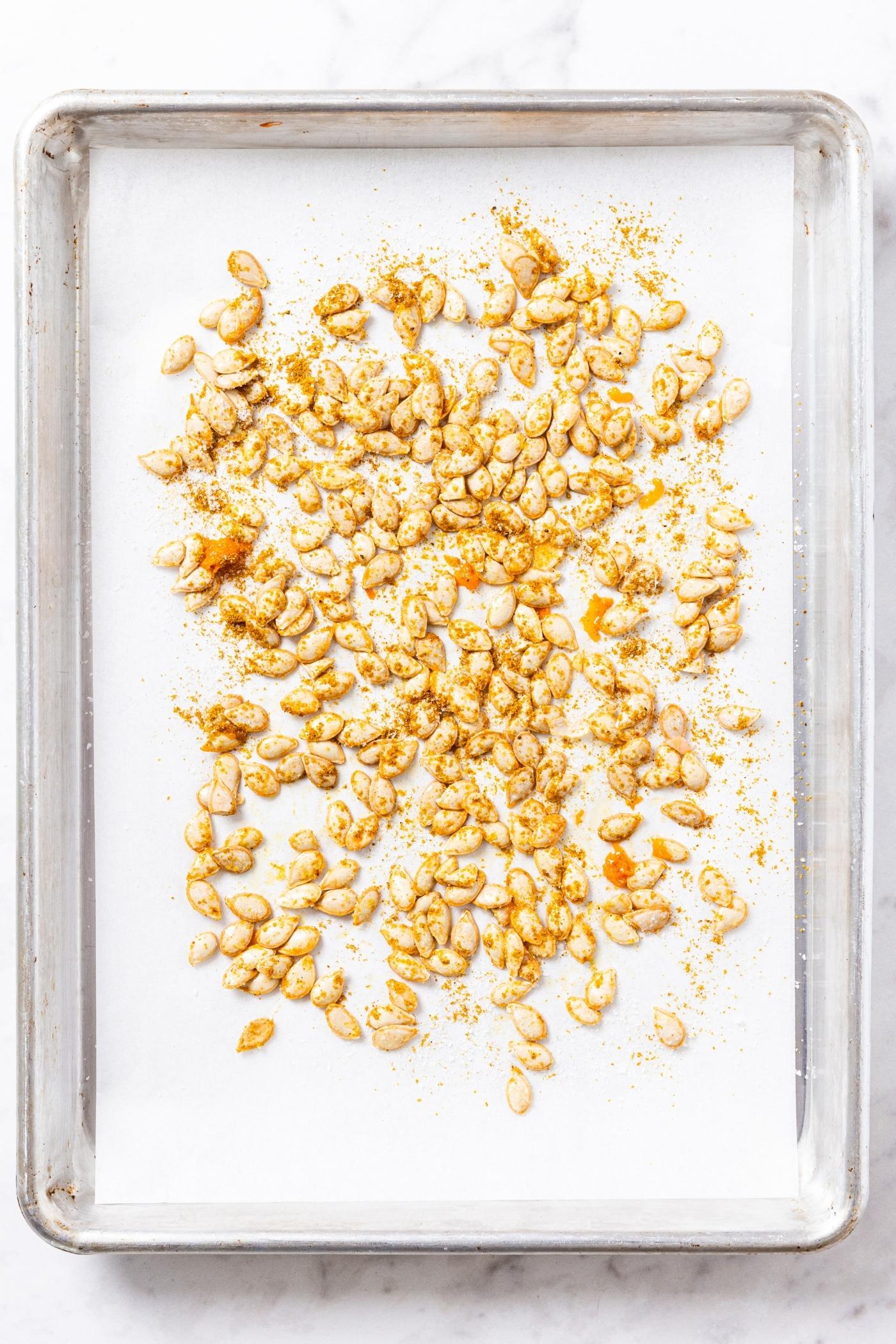 Metal baking pan with piece of white parchment paper with squash seeds spread out in even layer sprinkled with curry powder.