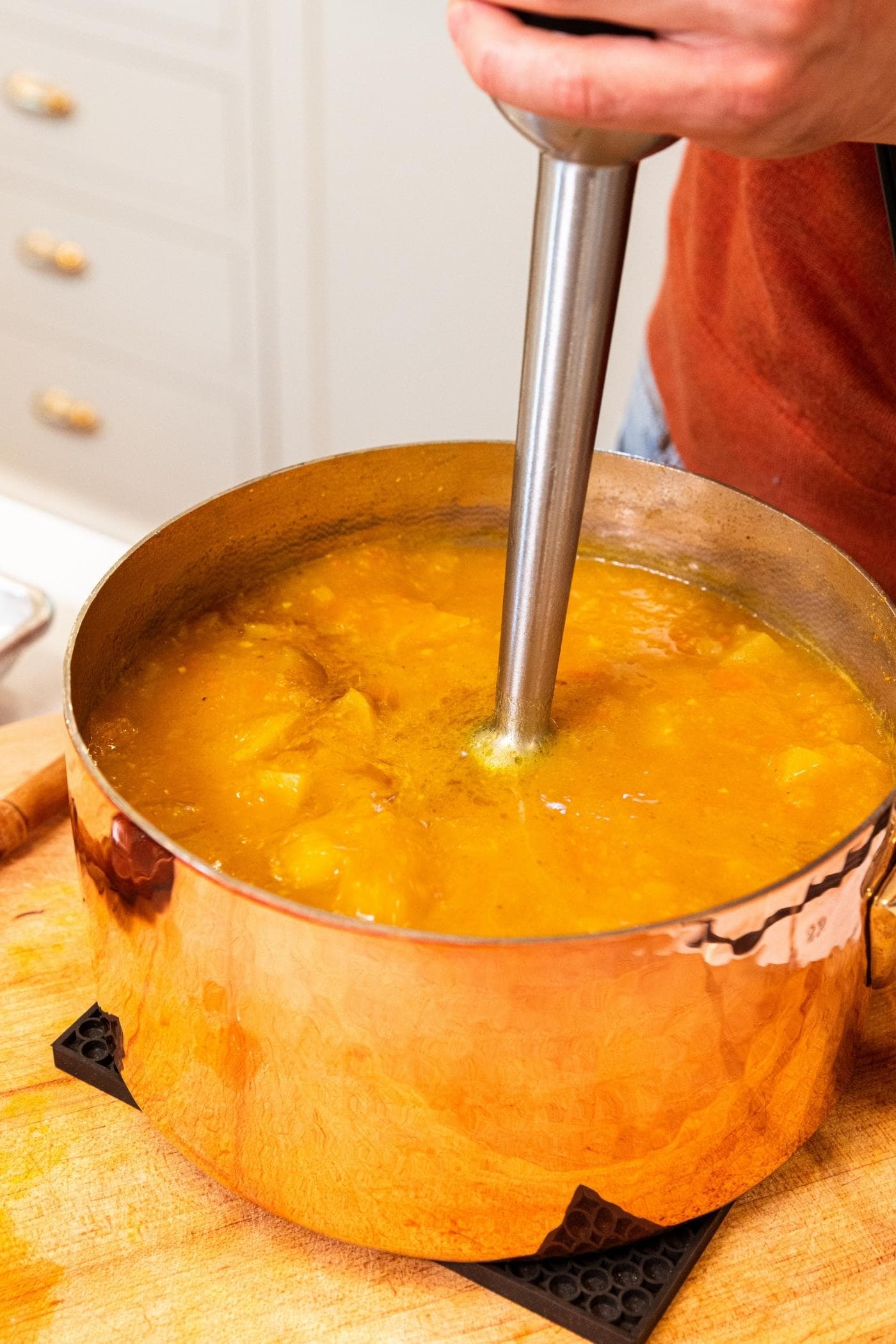 Copper pot sitting on black cooling hotpad on wooden cutting board with immersion blender in middle of pot blending the soup in pot together.
