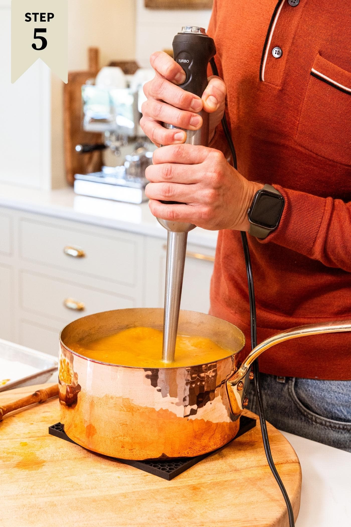 Two hands holding immersion blender blending butternut squash soup sitting in copper pot on wooden cutting board on white countertop.