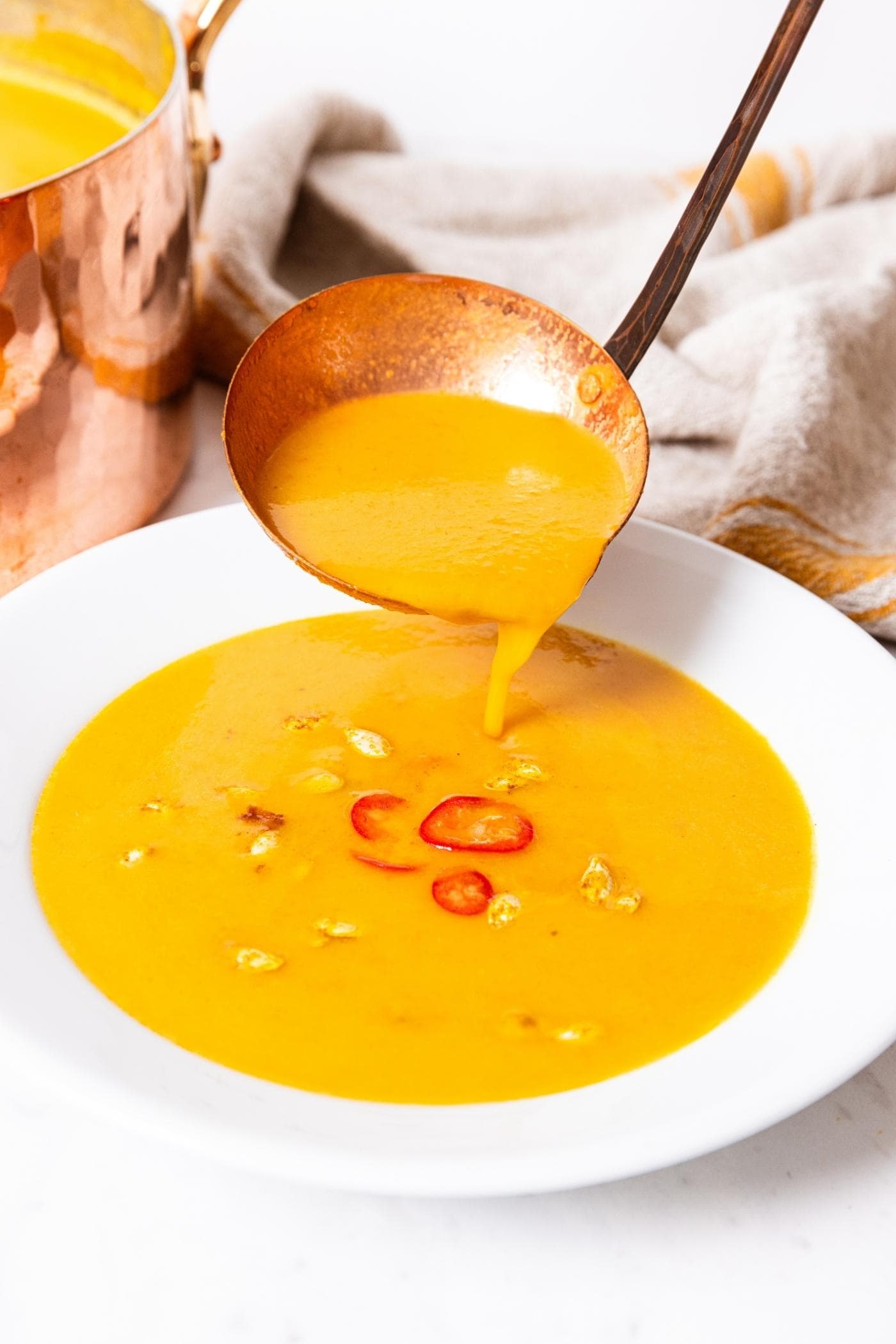 Copper ladle pouring serving of soup into a white bowl on marble surface with a few pieces of red pepper and squash seeds floating on top.