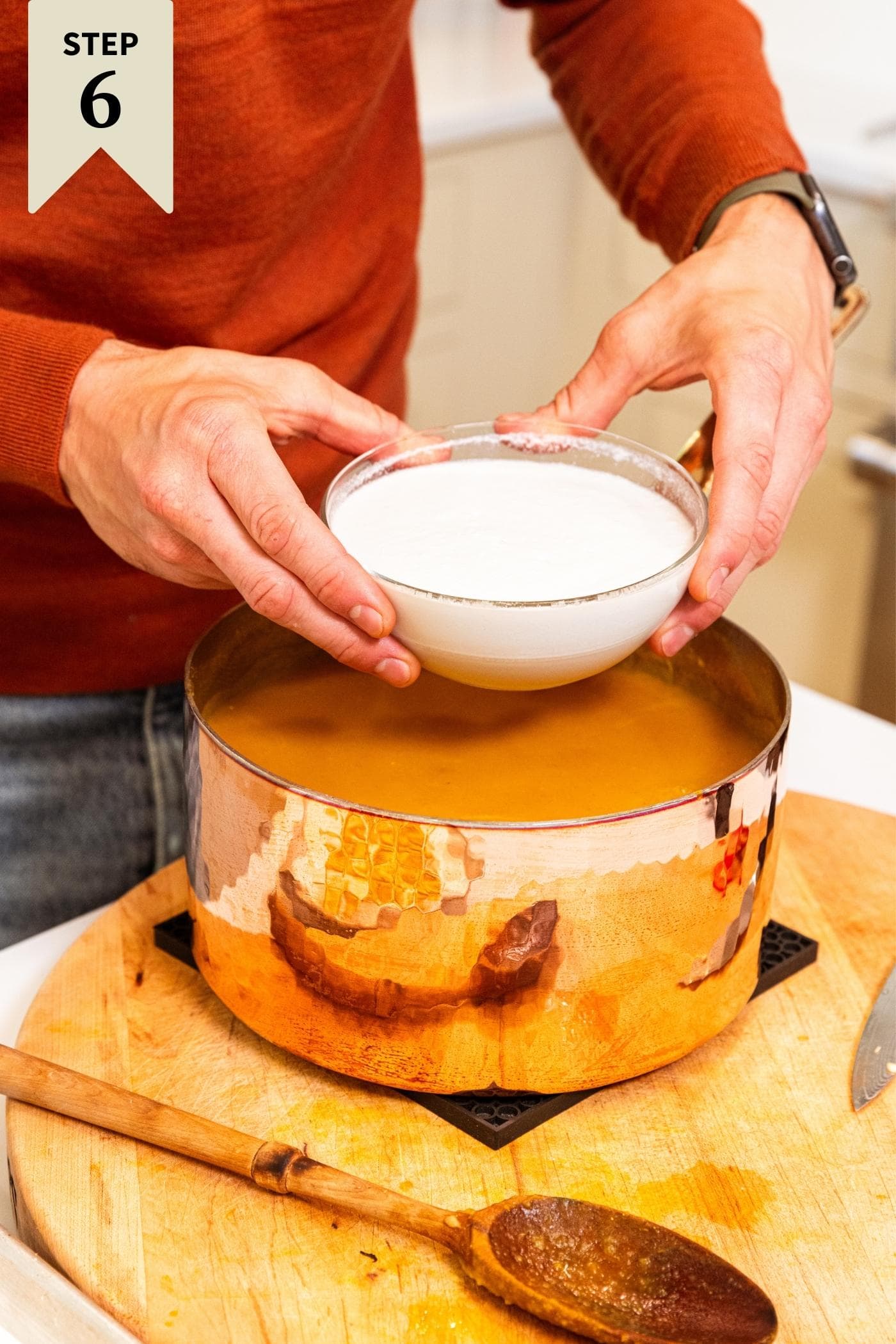 Two hands holding glass bowl filled with coconut milk pouring into a copper pot filled with butternut squash soup on wood cutting board.
