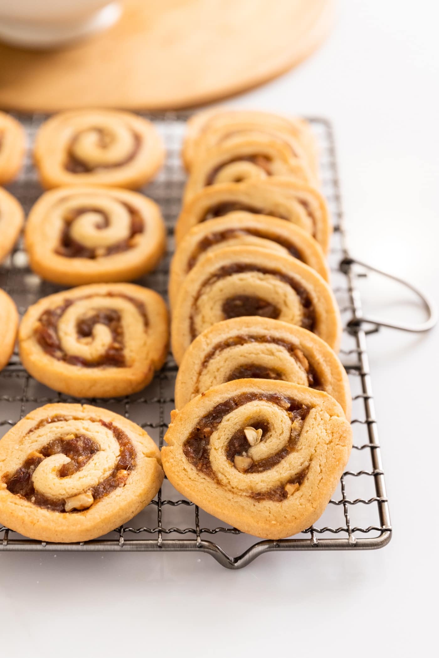 Date nut pinwheel cookies stacked against one another sitting on cooling rack with wood board and white bowl in background
