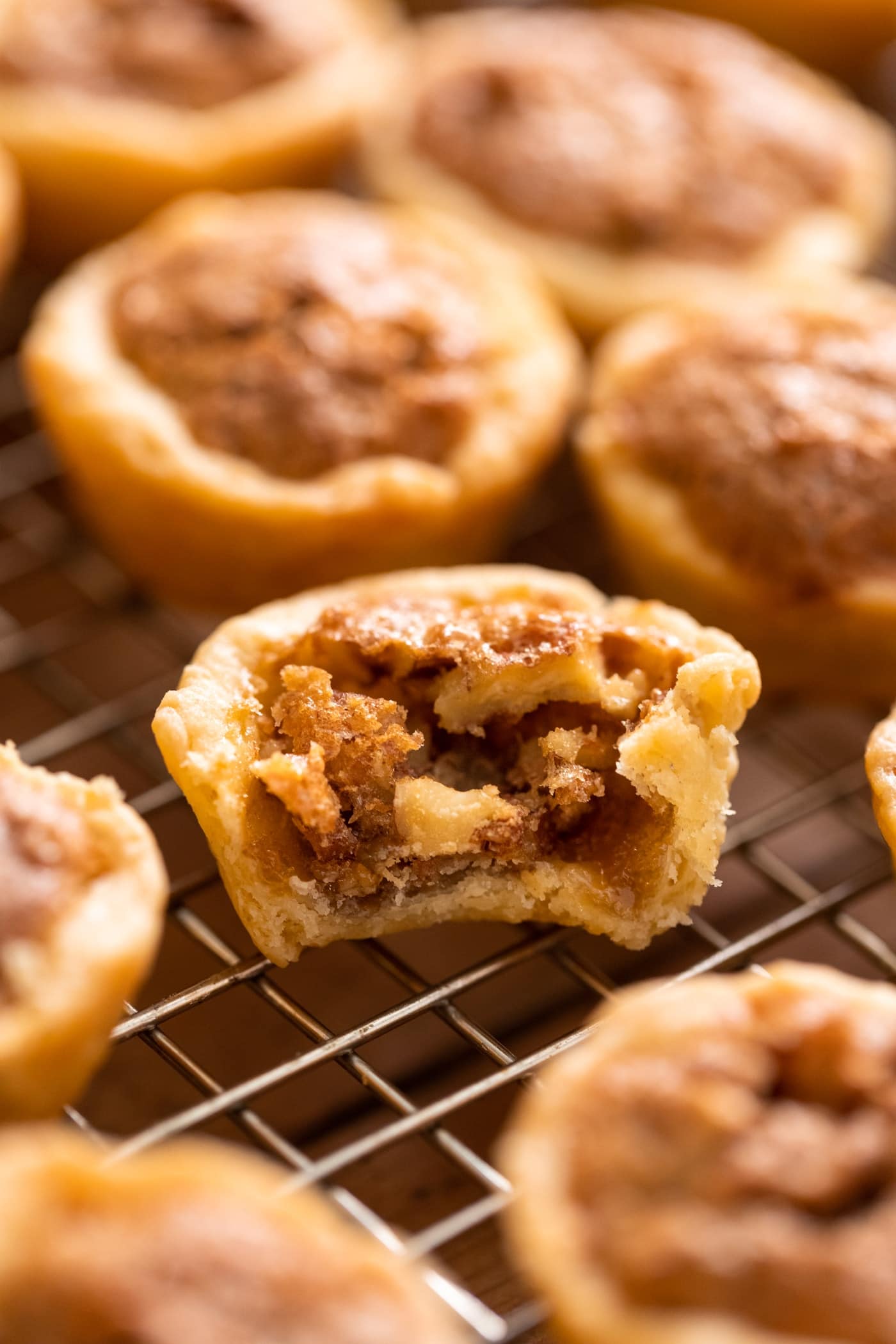 Small pecan tassie sitting on cooling rack with bite taken out with other pecan tassies sitting around