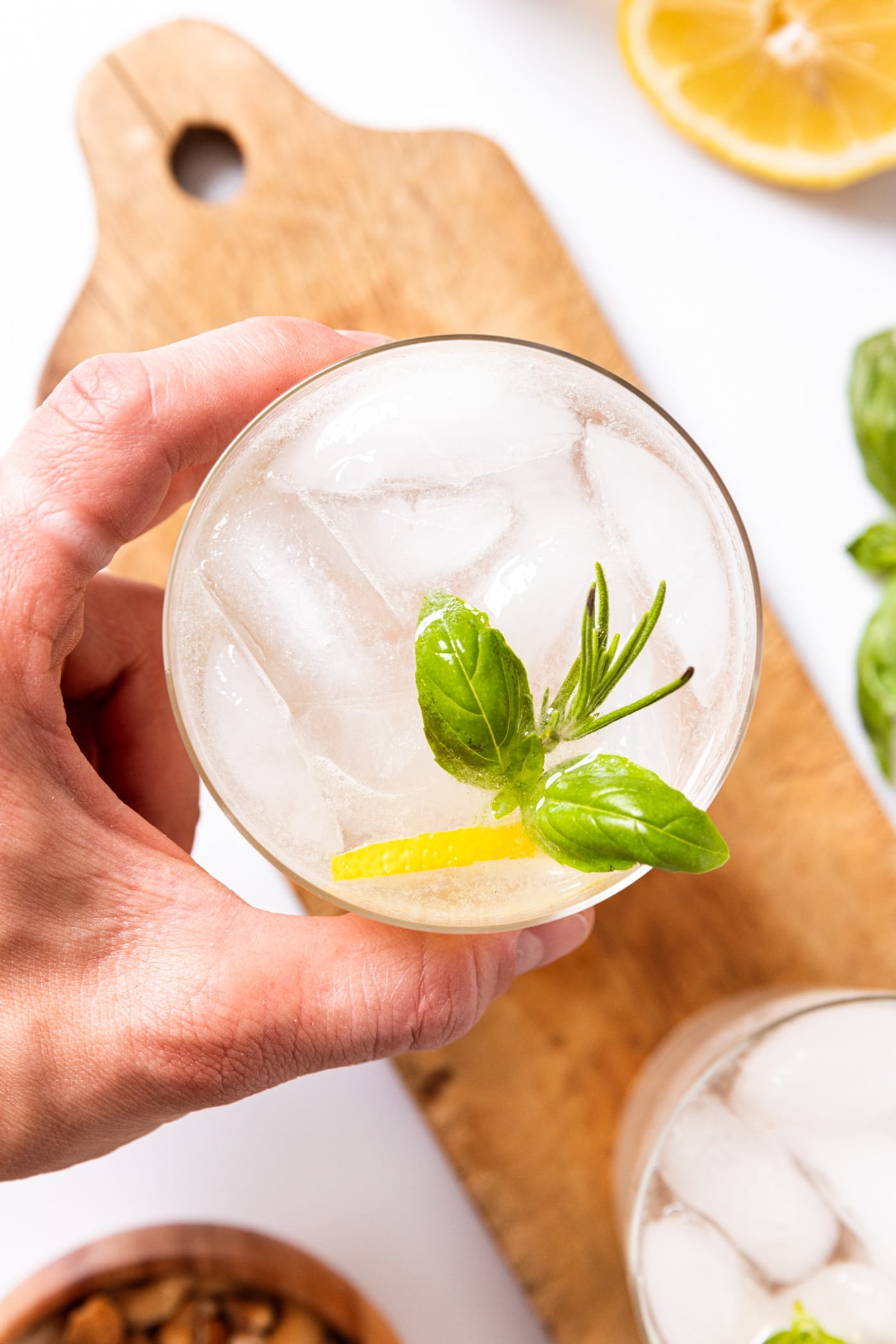 Glass filled with rosemary basil gin fizz with hand holding the glass with lemon wedge and herbs in glass with wood board underneath.