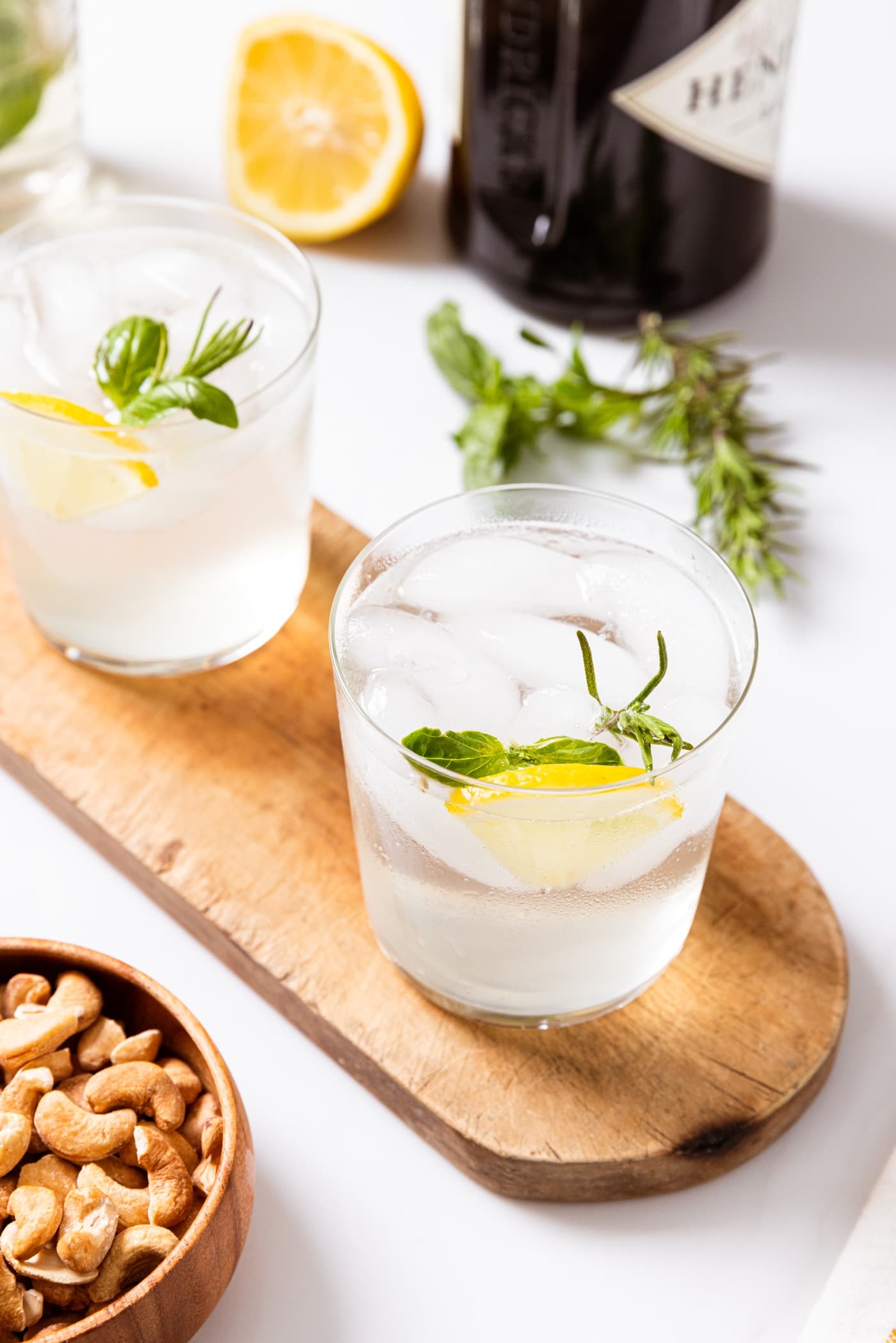 Two glasses filled with clear gin fizz liquid with lemon wedges and herbs stuck in glass sitting on wood board with small bowl of peanuts beside and alcohol container in background.