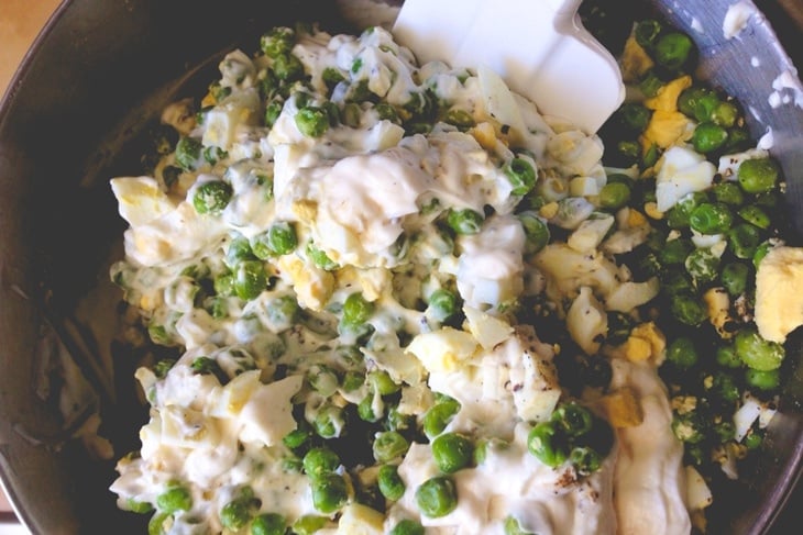 Mixing bowl filled with peas being mixed with sour cream and mayonnaise along with hardboiled eggs