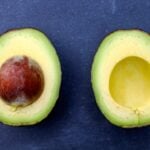 Two avocados one with a pit and the other without sitting on blue surface
