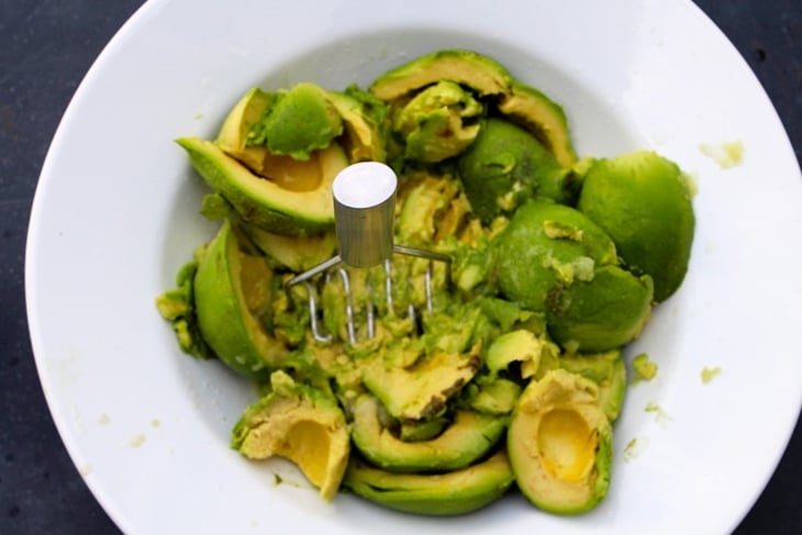 Potato masher mashing sliced avocados in white bowl
