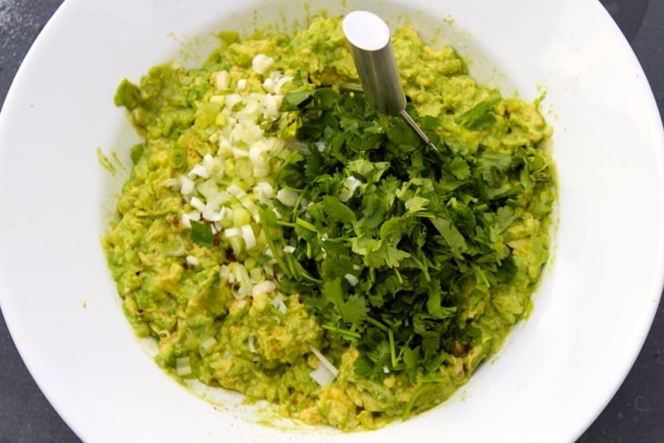 Avocado mixture with green onion and cilantro sitting in white bowl with potato masher
