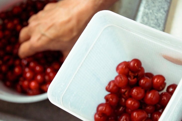 Hand placing washed and pitted sour cherries into plastic freezer-safe containers from a large bowl in a sink.