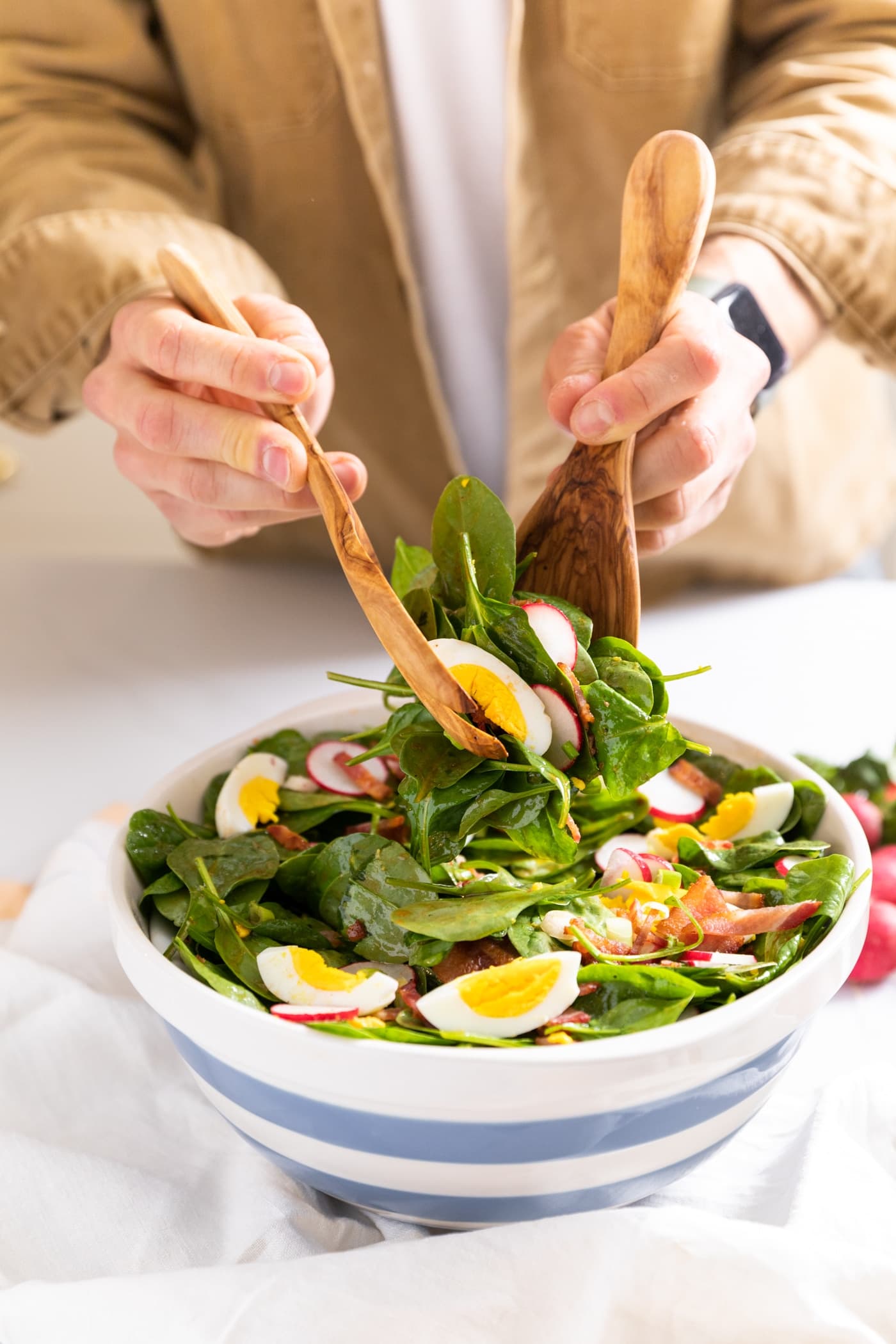 Two hands holding wooden serving spoons taking serving of spinach salad from large white bowl with blue stripes