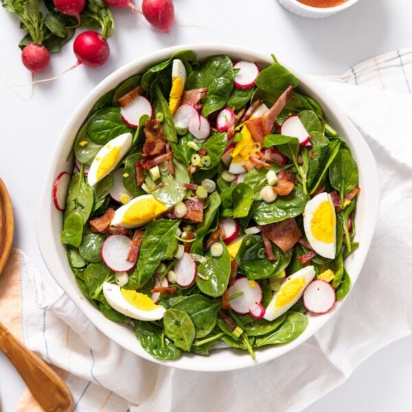 Top down view of white bowl filled with salad and topped with eggs, radish, and bacon with serving spoons and radish sitting on white countertop