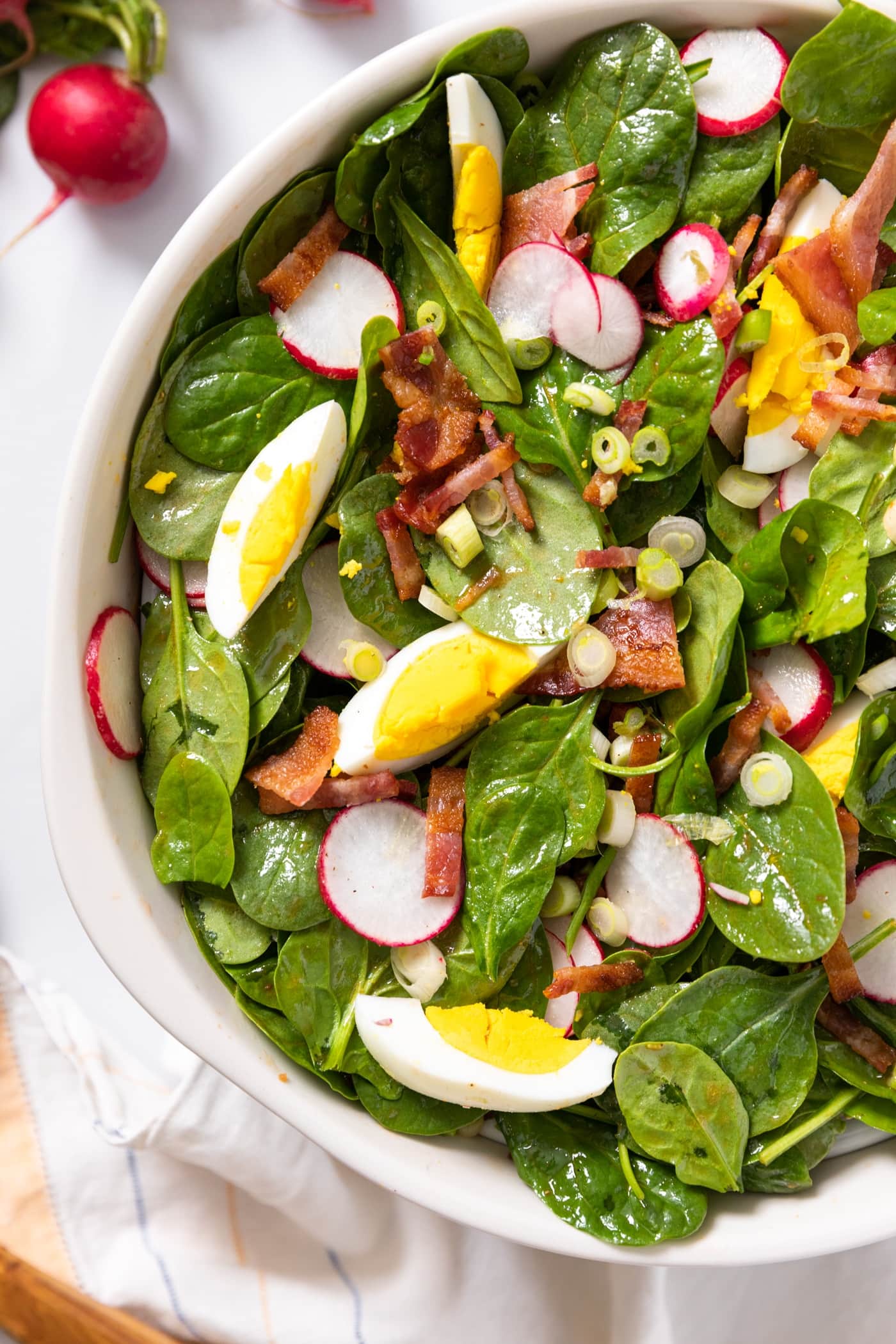 Top down view of white bowl filled with spinach leaves topped with hardboiled egg, bacon, and radish