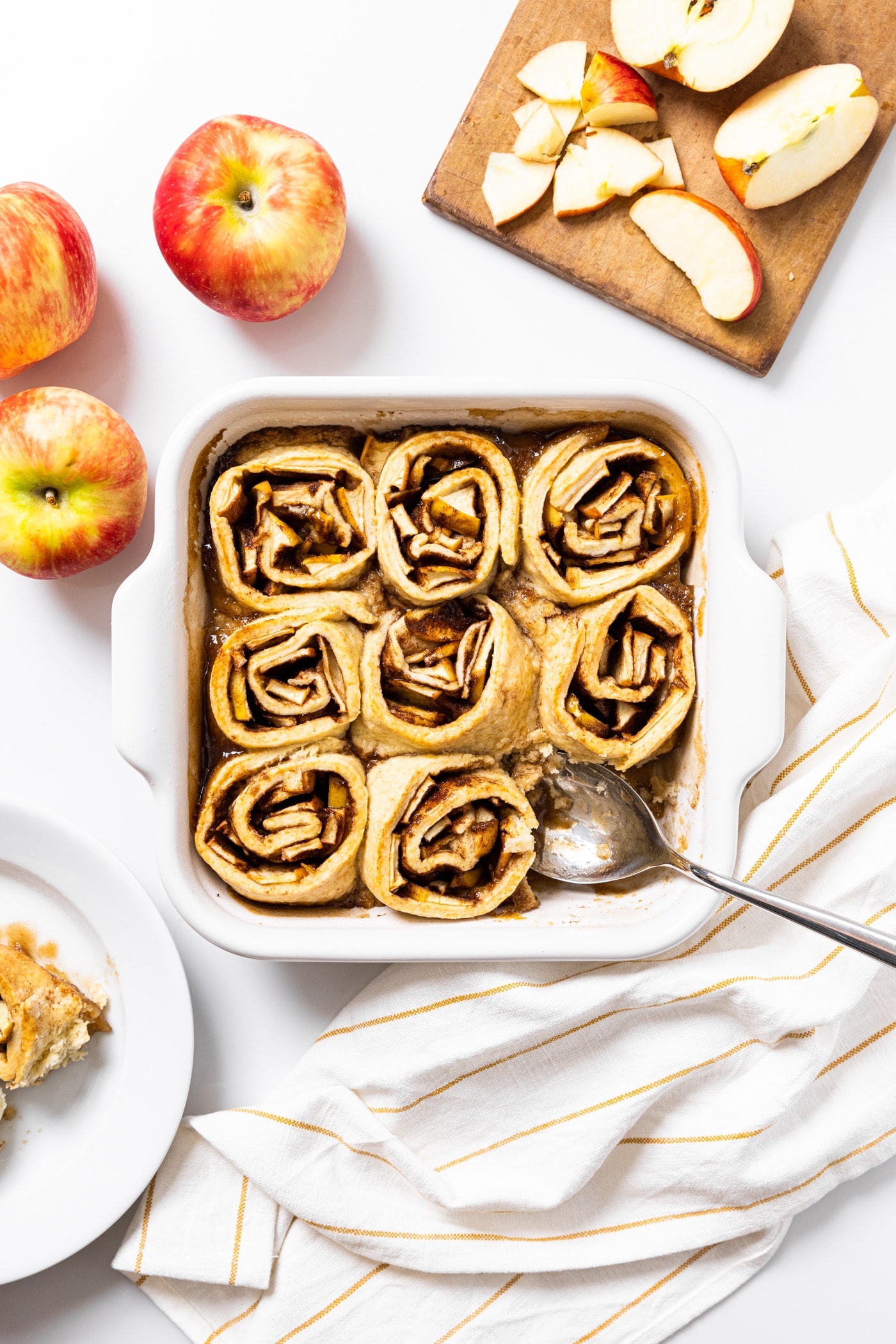 Further away top down view of white baking pan filled with cinnamon filled apple dumpling rolls with apples and plate filled with one roll