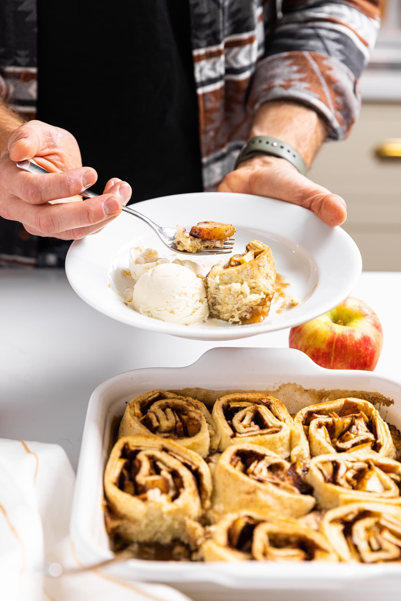 White plate being held by hand with a fork full of apple from one slice of apple dumpling along with a scoop of white ice cream