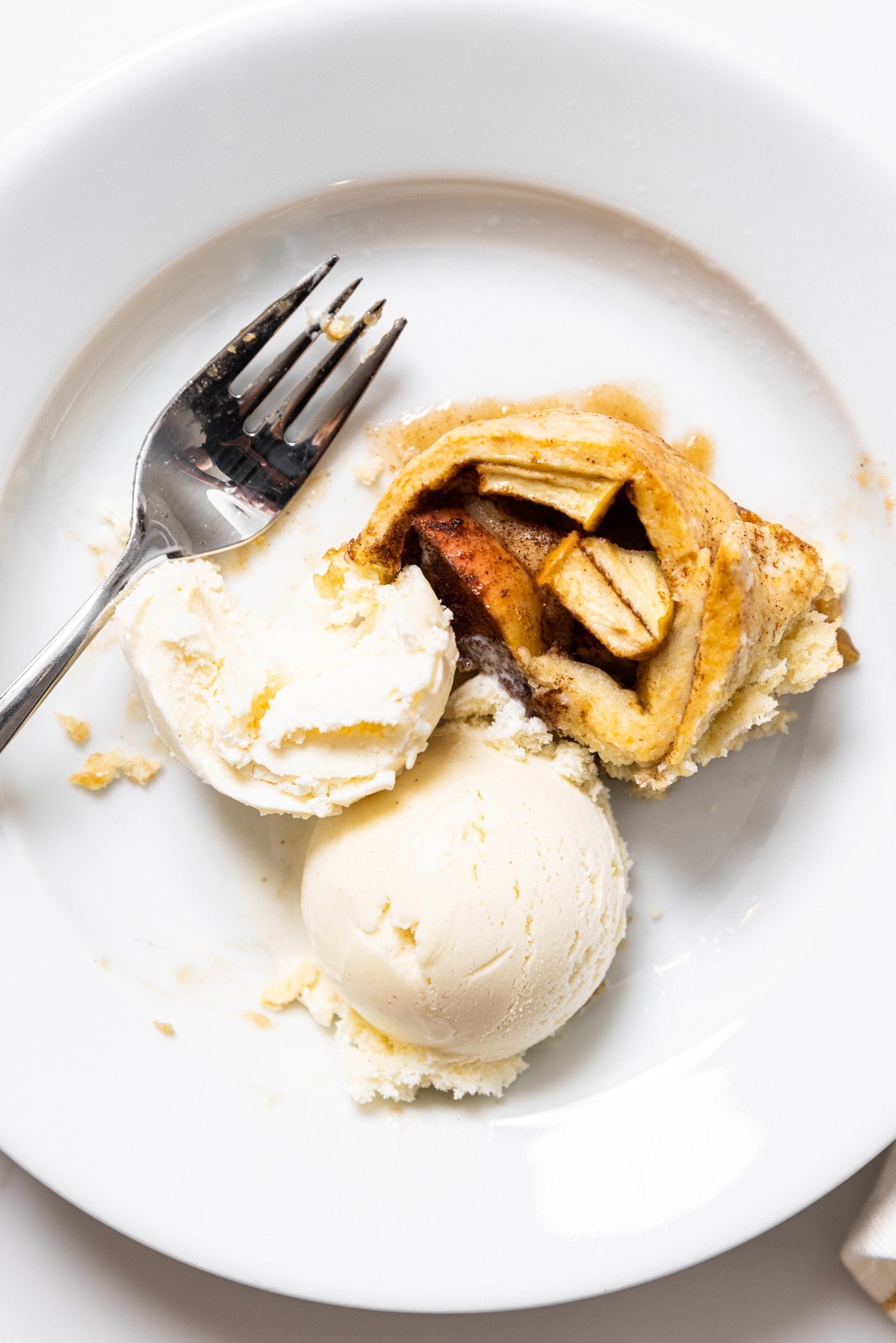Top down view of white plate filled with two scoops of white ice cream and partially eaten apple dumpling roll