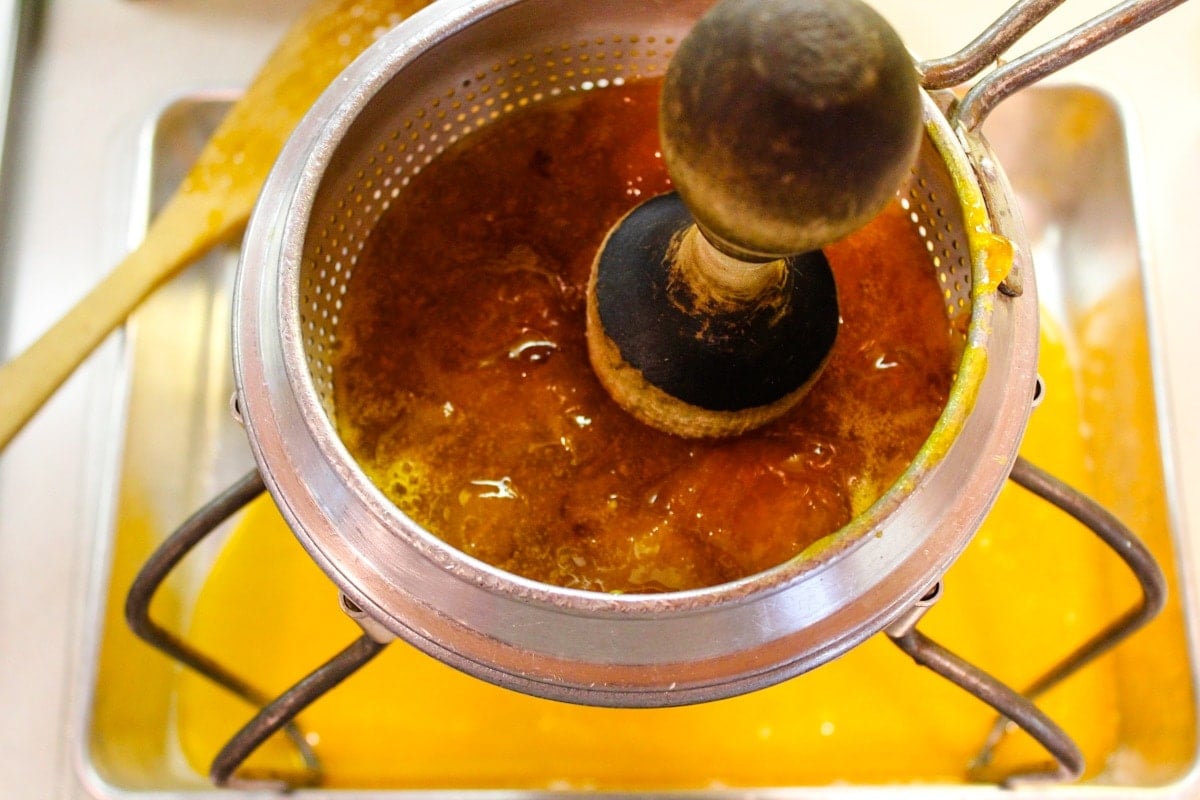 Apricot butter mixture sitting in a conical strainer with wooden pestle all over a 9x13 pan catching all liquid