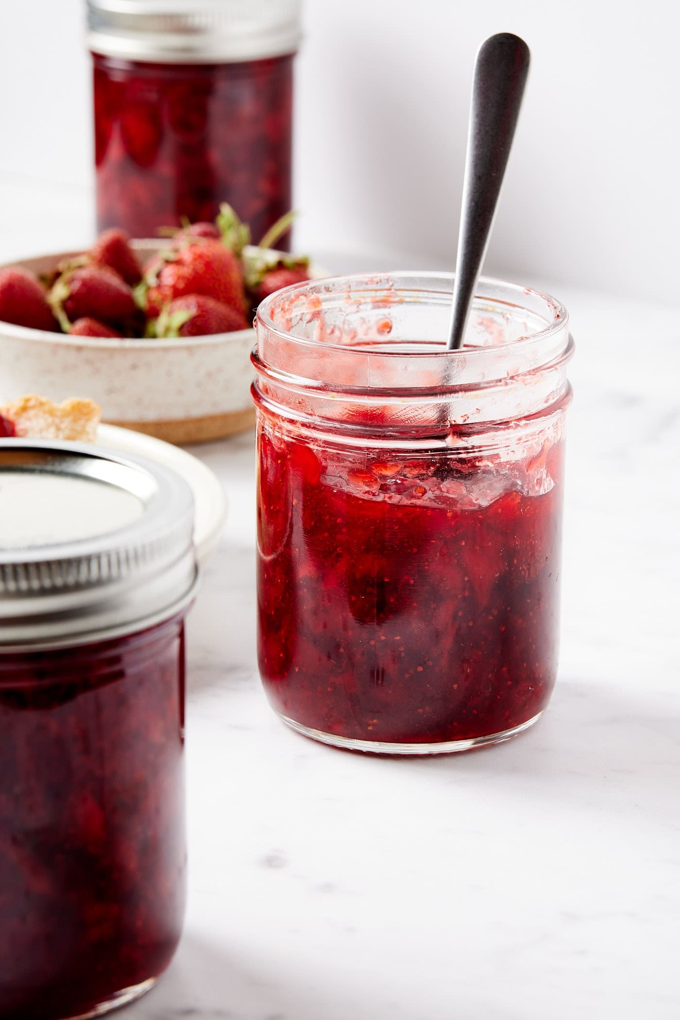 Jar filled with strawberry jam with spoon inside on white marble surface with more jam jars around.