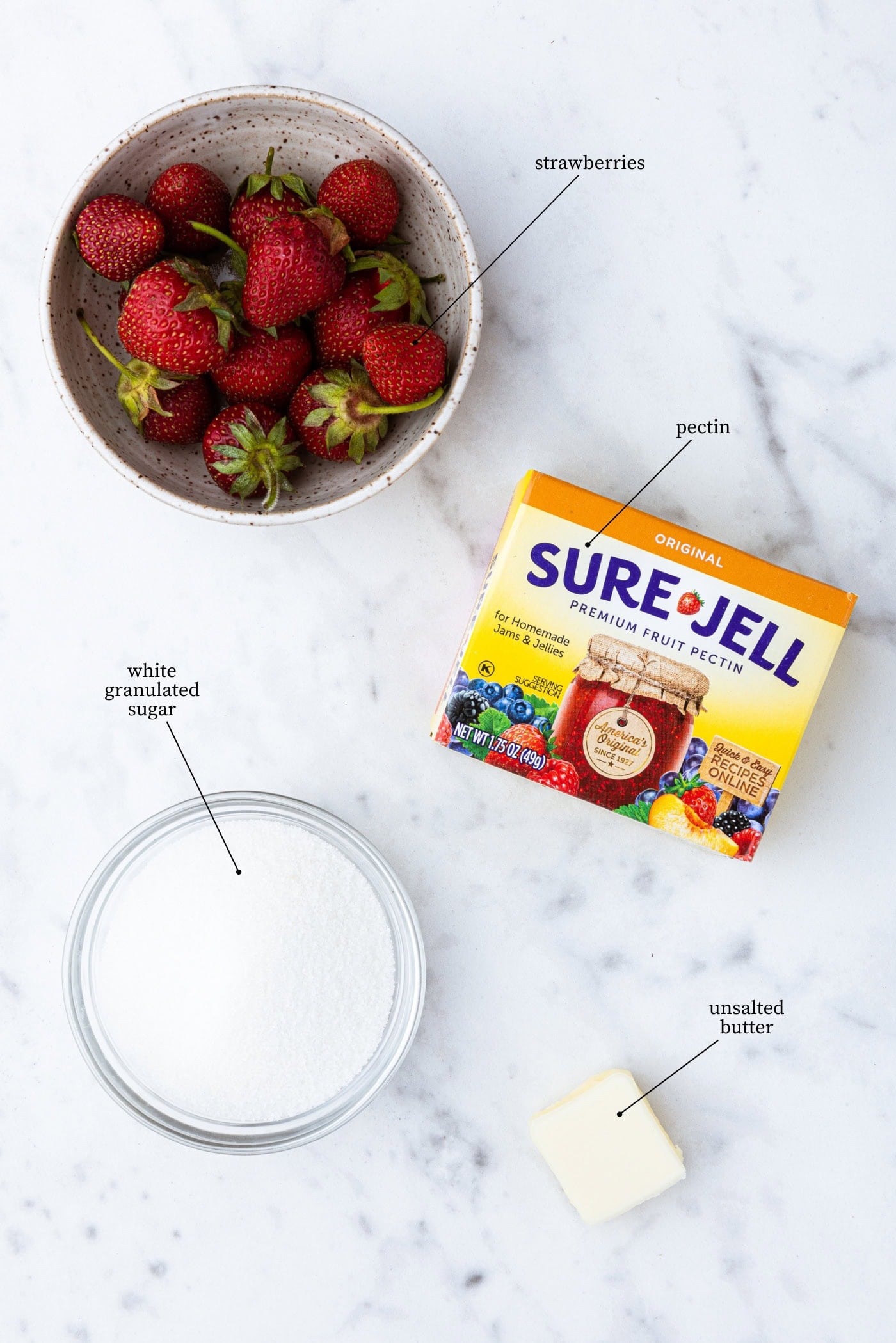 White marble surface with ingredients needed for strawberry jam including sugar, strawberries, Sure-Jell, and butter.