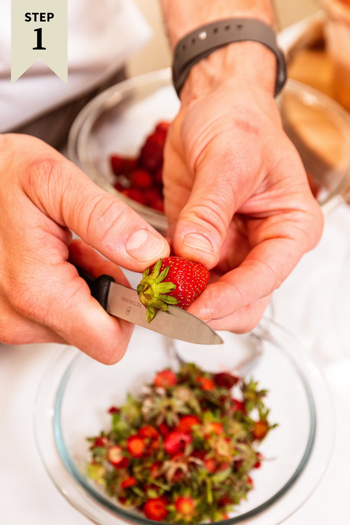 Hand slicing the top off of strawberries with small bowl filled with discard on top.
