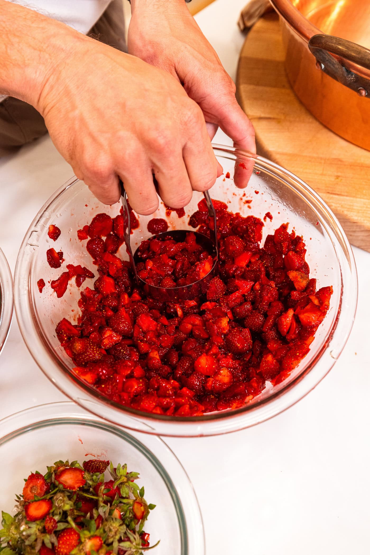 Hand using masher to chop raw strawberries in a large glass bowl.