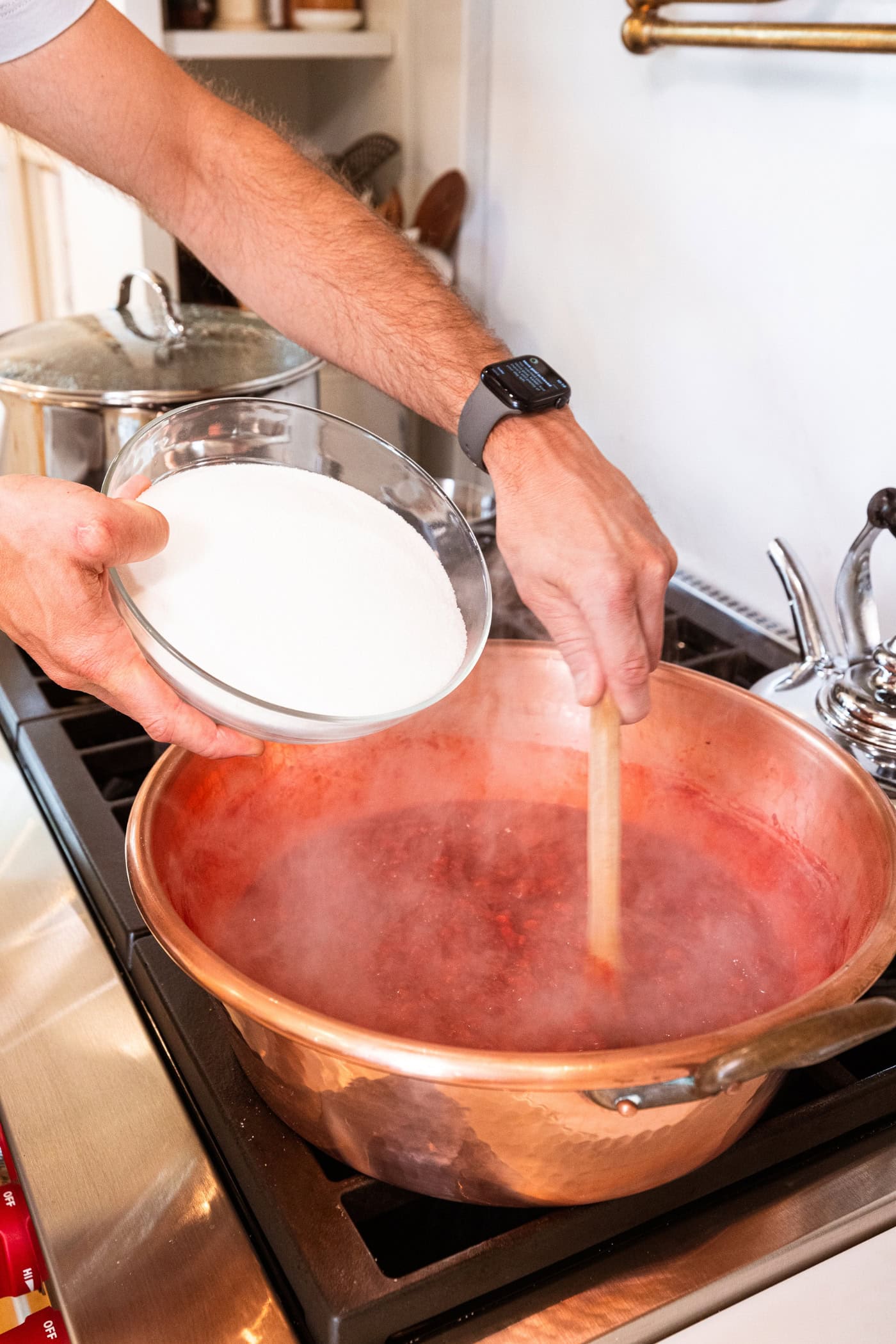 Hand about to pour sugar from a bowl into a copper pot filled with cooking strawberries for strawberry jam recipe.