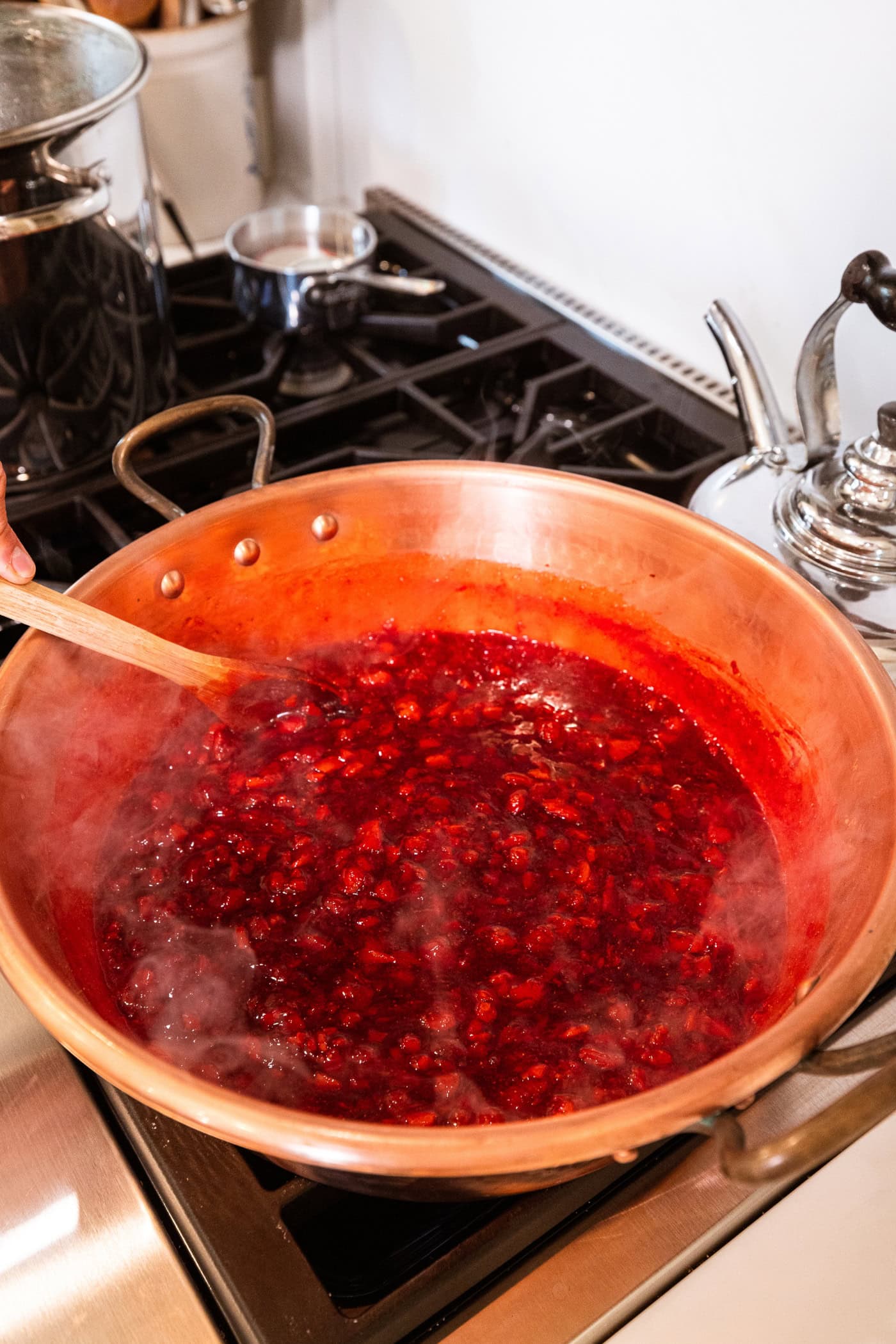 Large jam pan on the stovetop with cooking sugar and strawberry mixture with steam coming off the top.