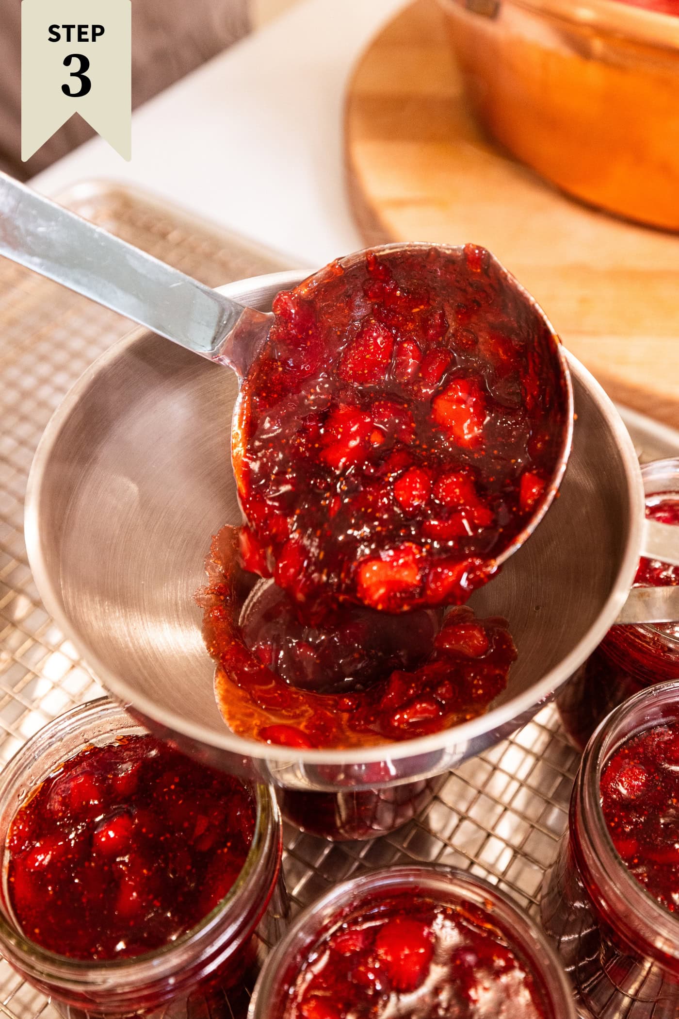 Ladle pouring strawberry jam through a funnel into jam jars filled with jam.