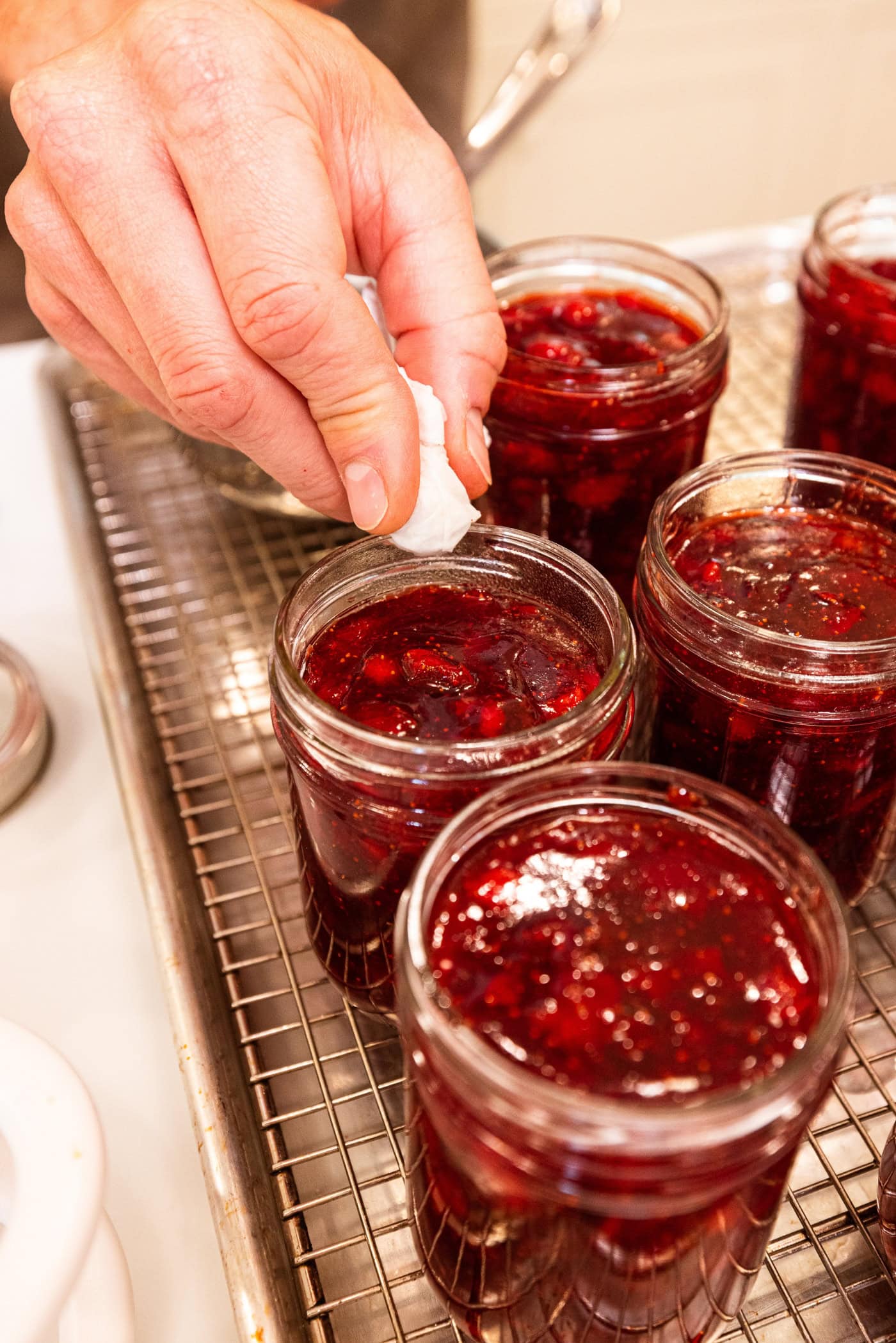 Hand using small piece of paper towel to wipe off the rim of a jam jar filled with jam.