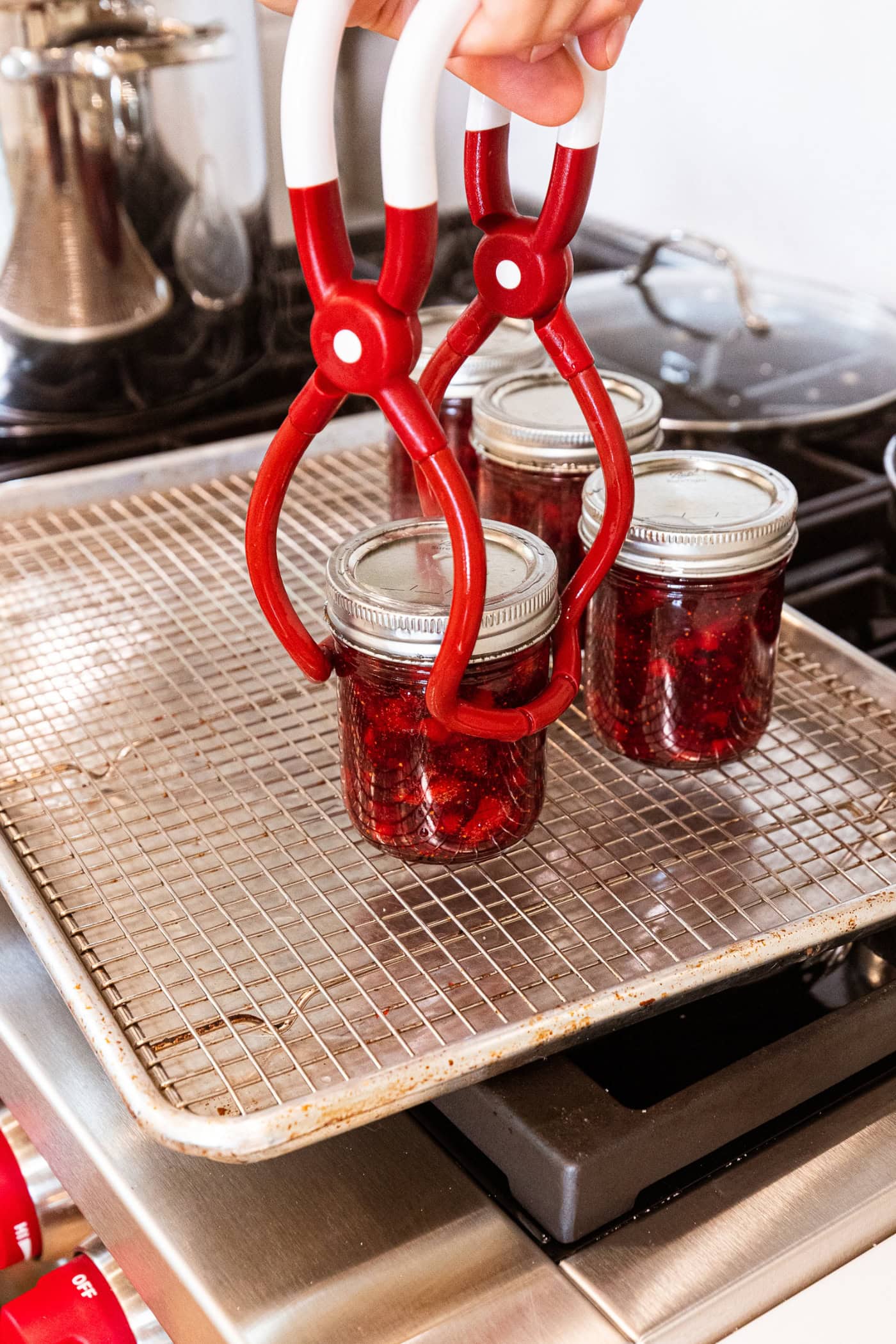 Jar lifter setting jam jar filled with strawberry jam on a wire rack to cool after coming out of the canner.