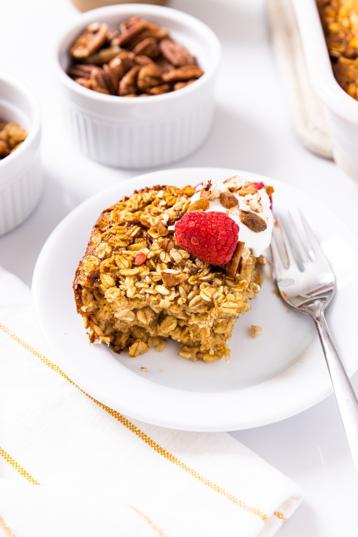 Square piece of golden brown baked oatmeal sitting on white plate with raspberry and Greek yogurt on the side with nuts in white container in background
