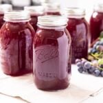 Glass jars filled dark purple canned grape juice concentrate sitting on white countertop with grapes sitting behind