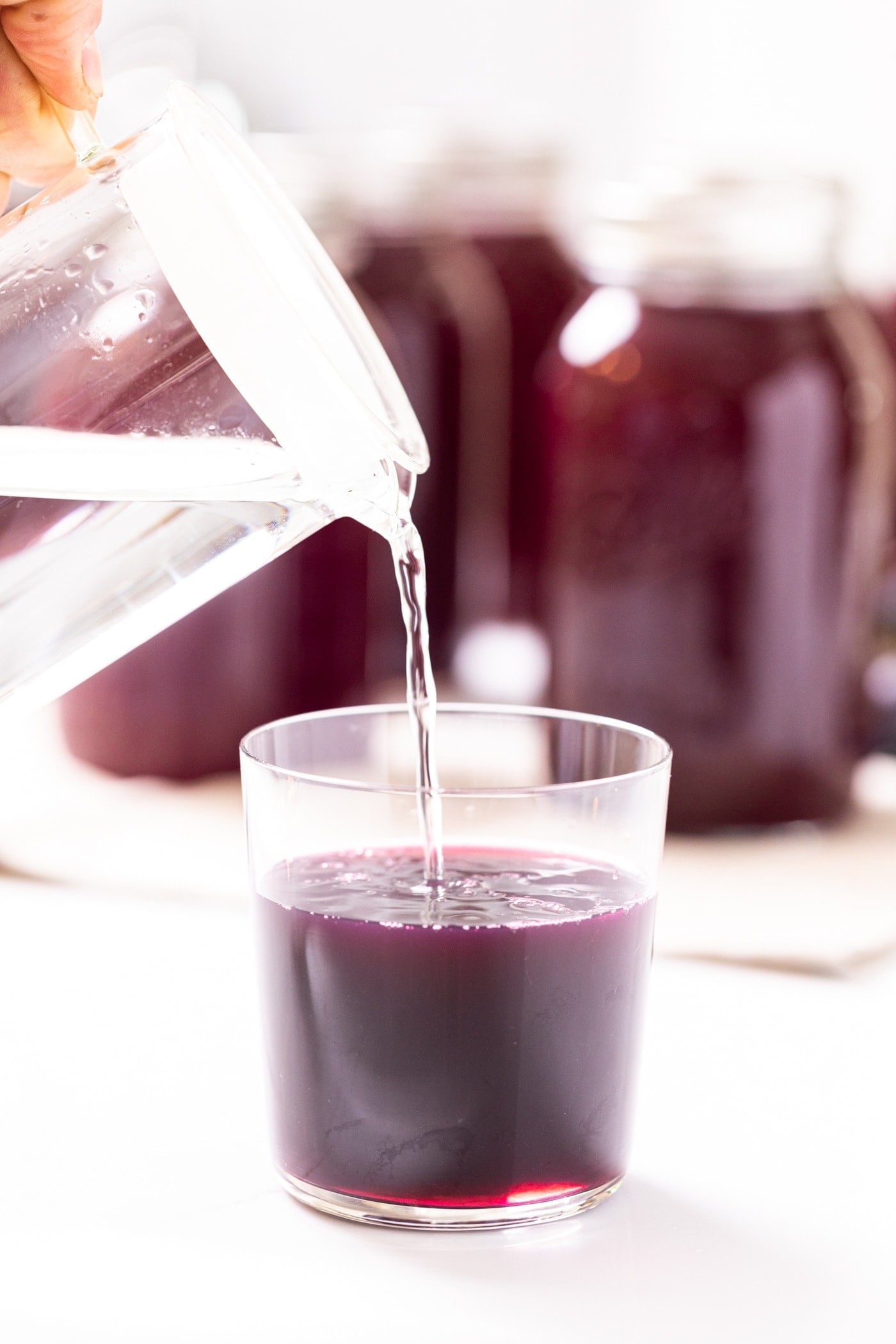 Glass filled with dark purple grape juice concentrate being filled from glass pitcher with glass jars in background