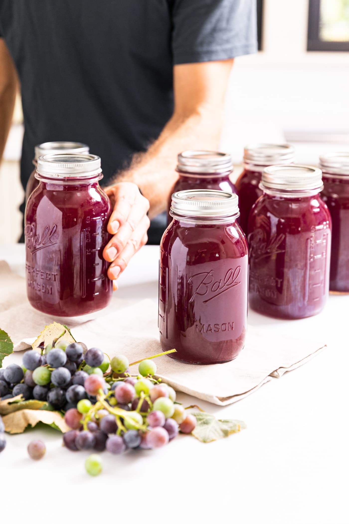 Hand holding glass jar filled with dark purple grape juice concentrate sitting on white countertop with grapes in front