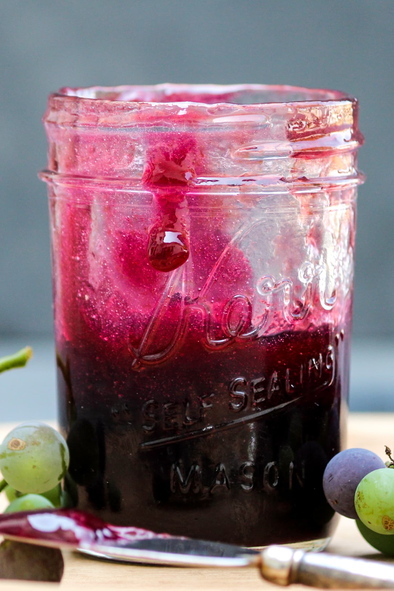 Single bead of grape butter running down the side of jam jar filled with purple grape butter on a wood cutting board with serving knife and grapes to the side on a piece of gray slate