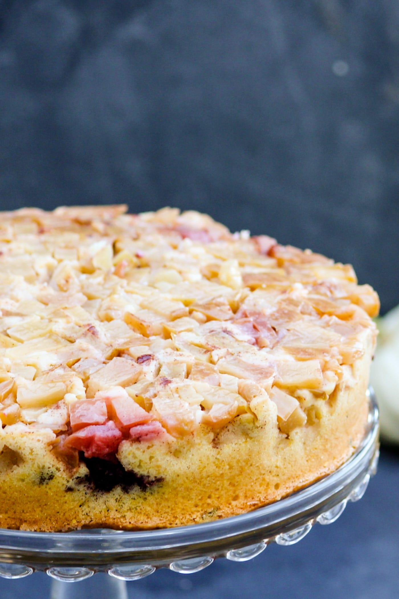 Harvest apple cake topped with apples and blackberries sitting on a cake platter with small white pumpkin in background with a gray slate surface