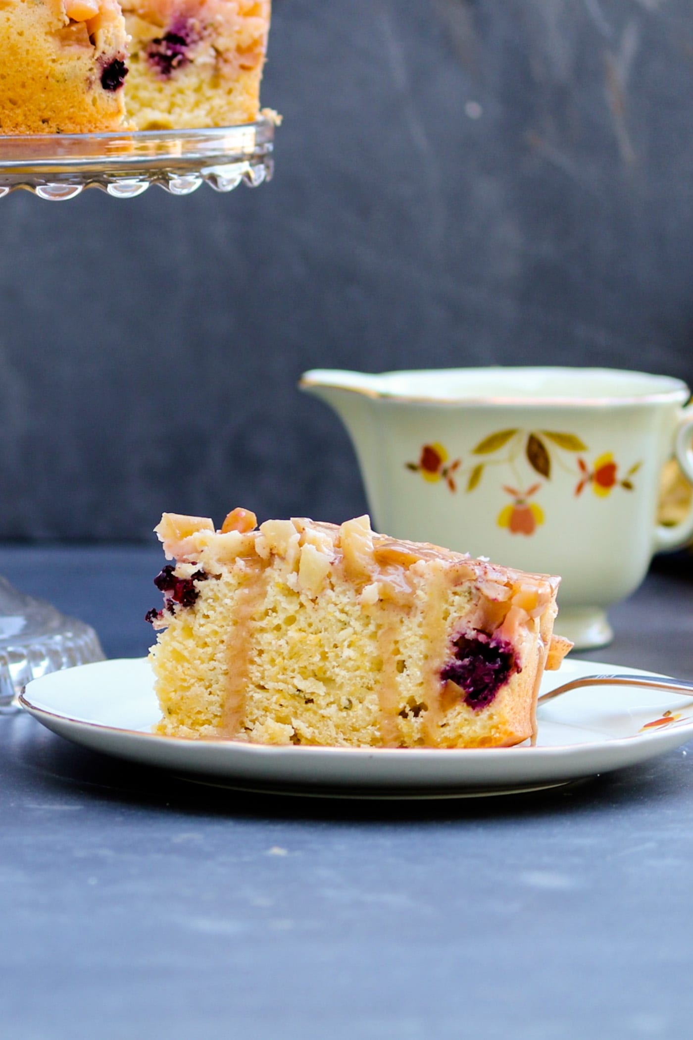 Piece of harvest apple cake sitting on a white plate with glaze drizzled over top with coffee cup in background on a gray slate surface