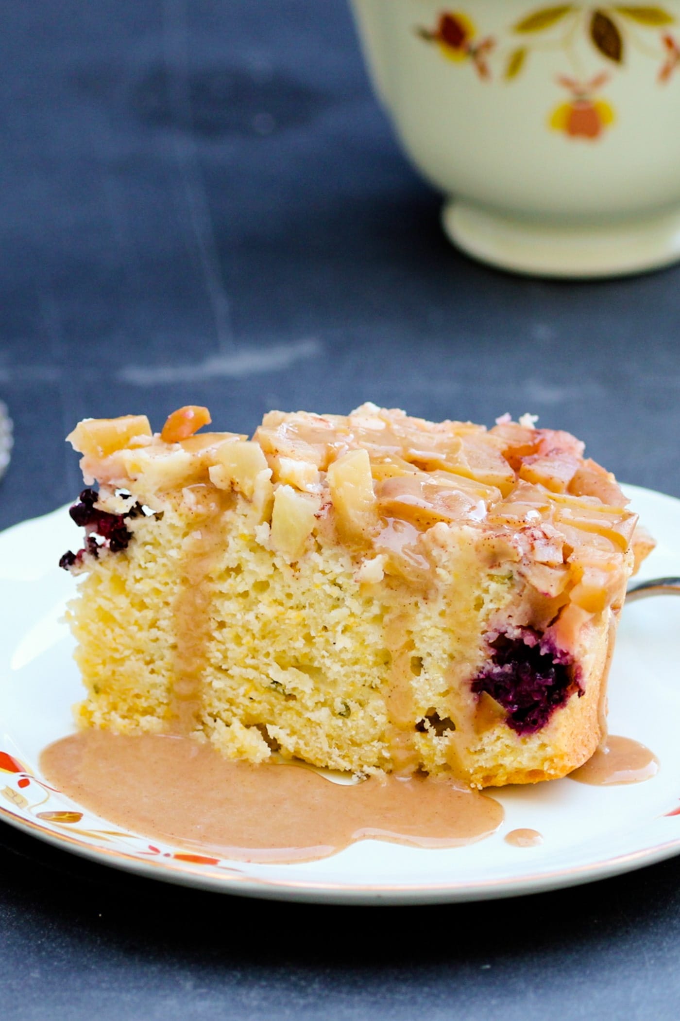 Piece of harvest apple cake sitting on a white plate with glaze drizzled over top with coffee cup in background on a gray slate surface